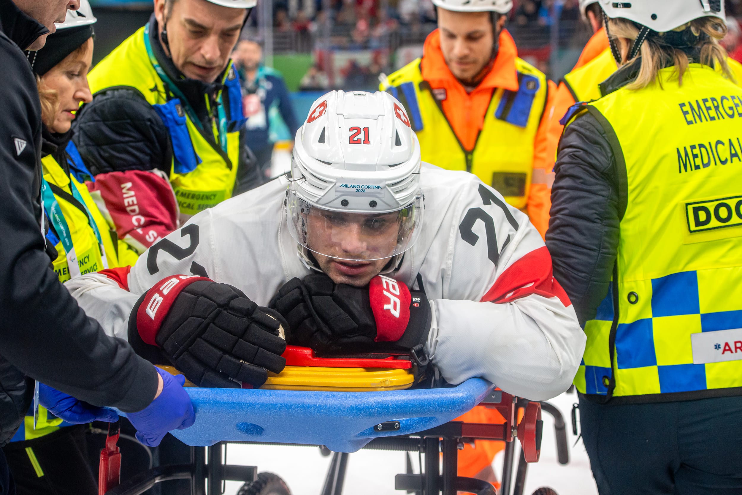 A hockey player wearing a white and red jersey and helmet lies facedown on a stretcher, surrounded by emergency medical personnel.