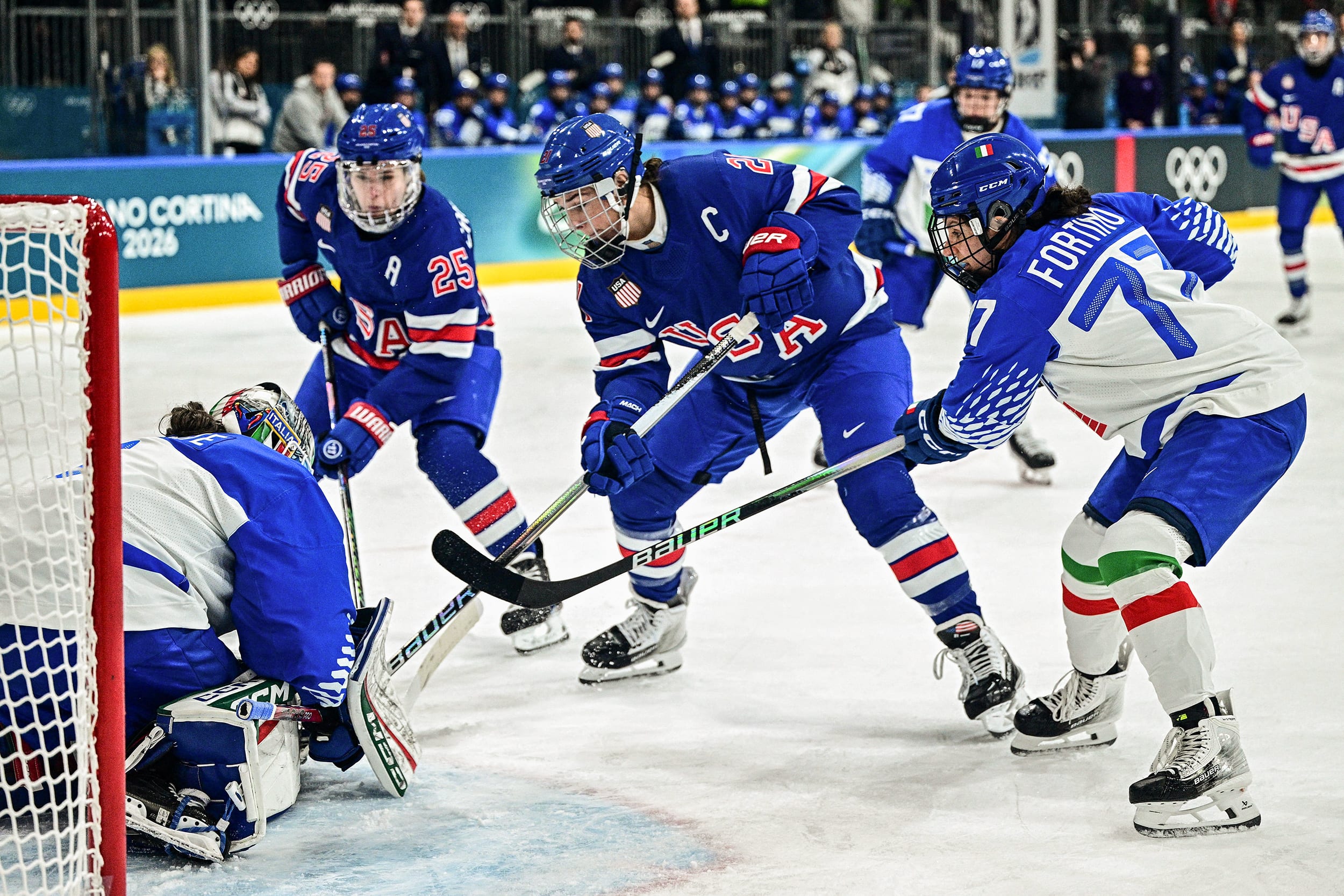 Hilary Knight is crouched in front of the opposing team's goalie, flanked by two other players.