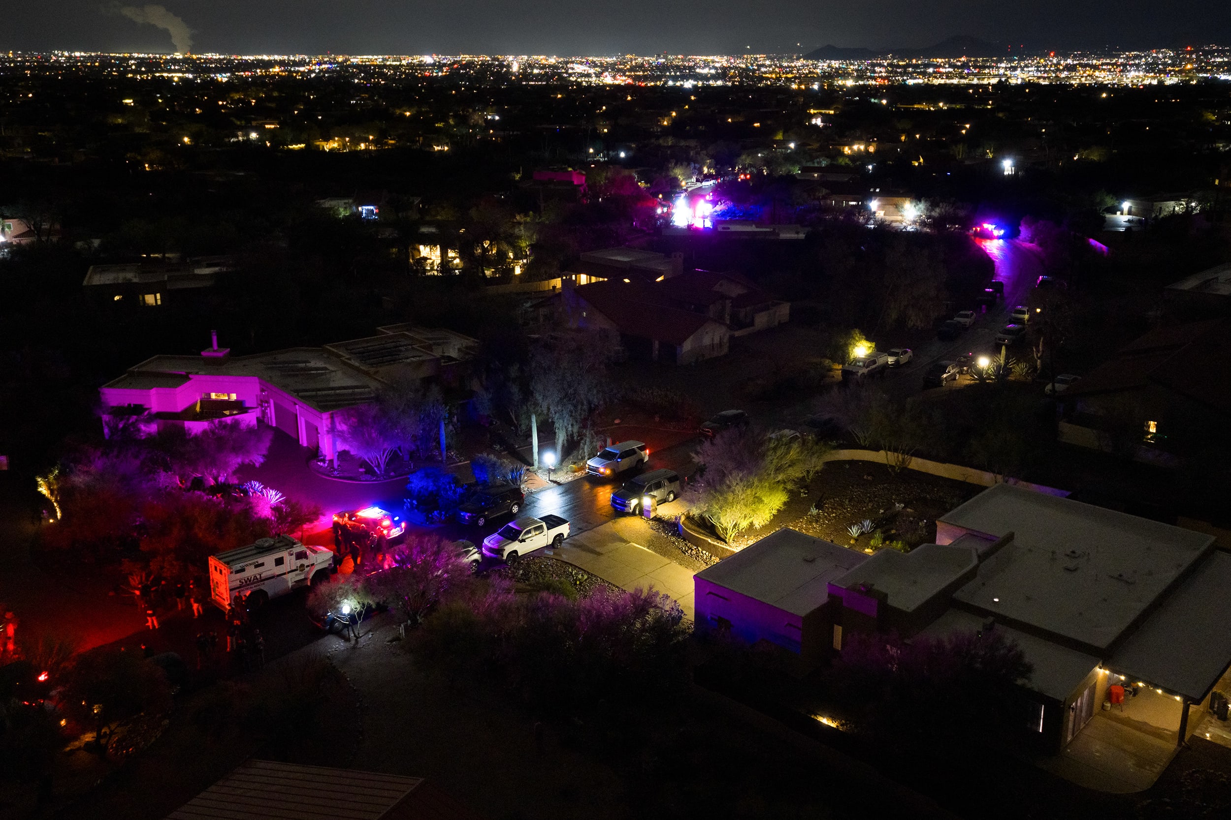 An aerial view shows dark residential streets partially illuminated by red, purple, and blue lights from law enforcement vehicle lights.