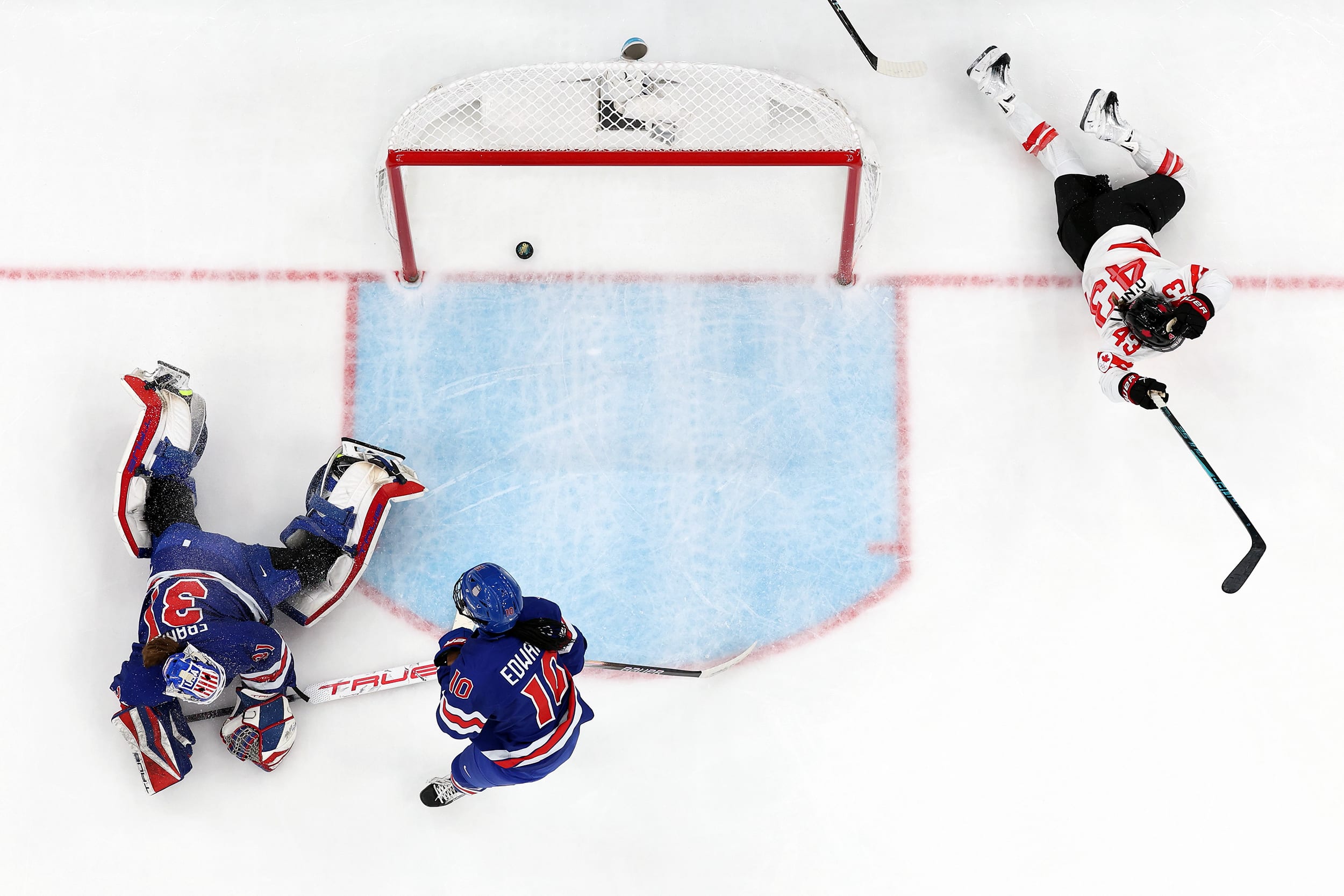 An overhead view of three ice hockey players surrounding a goal.