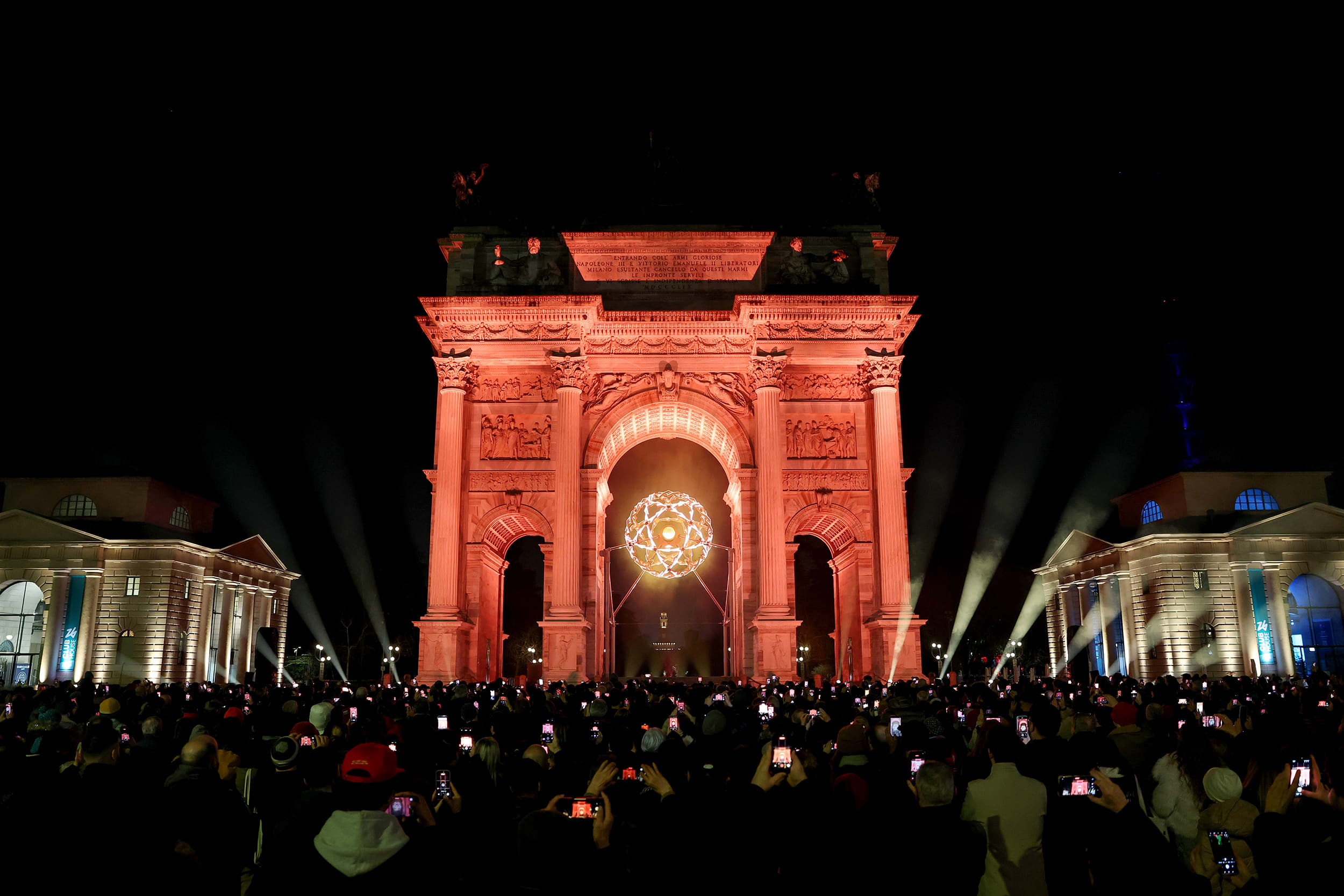 A glowing orb illuminates the stone arch where it is suspended above a large crowd of people holding mobile phones up.