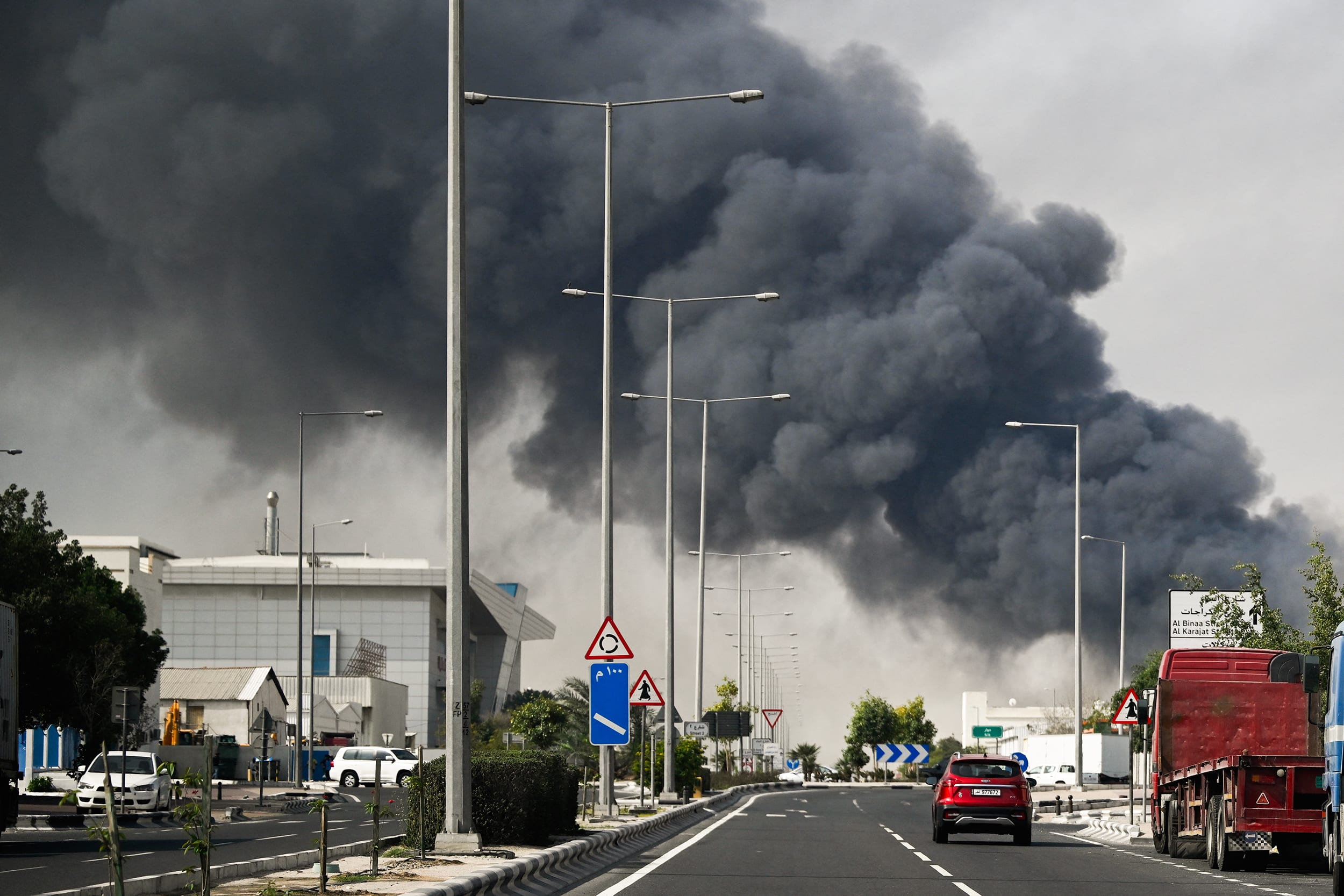 A huge cloud of thick black smoke rises above road traffic. The entire sky is gray.