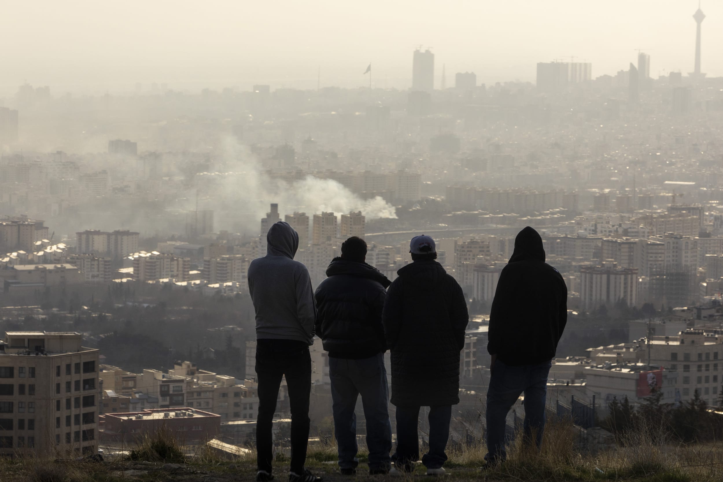 Men watch from a hillside as a plume of smoke rises after an explosion on March 2, 2026 in Tehran, Iran. 