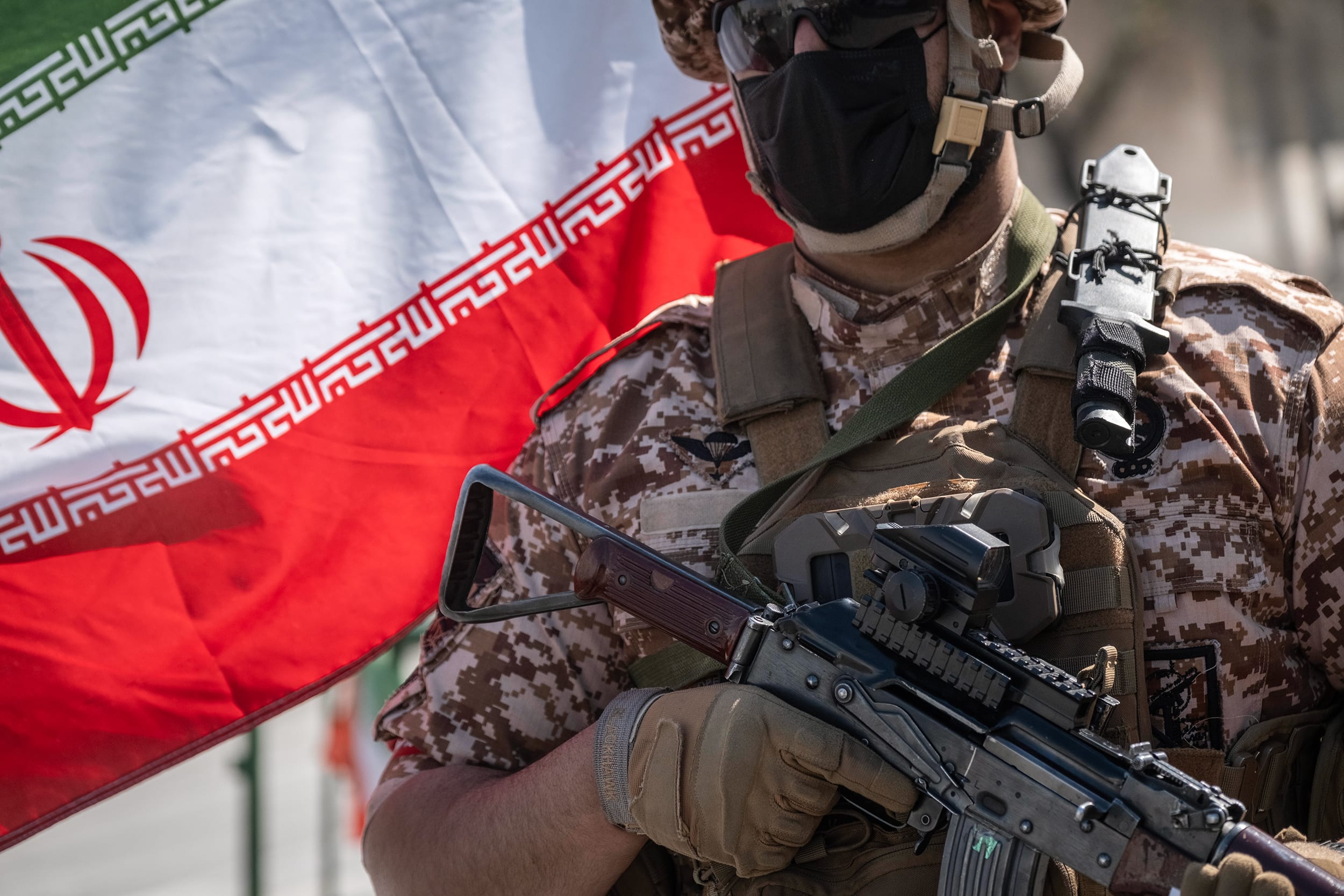 A soldier wearing a black surgical mask, sunglasses and fatigues holds an assault rifle in front of the Iranian flag.