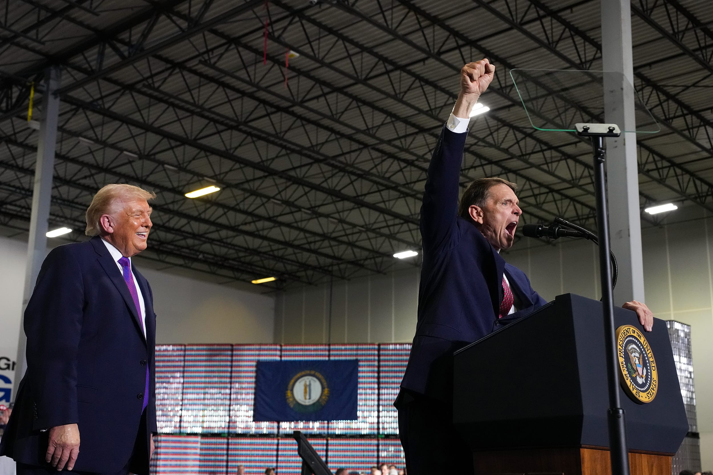 Donald Trump smiles behind Ed Gallrein, who is shouting at a lectern.