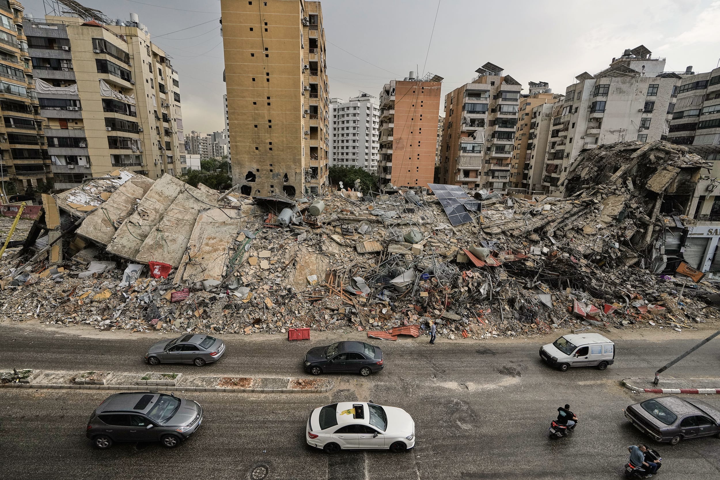 Heavily damaged buildings and rubble lie alongside a street with cars passing by.
