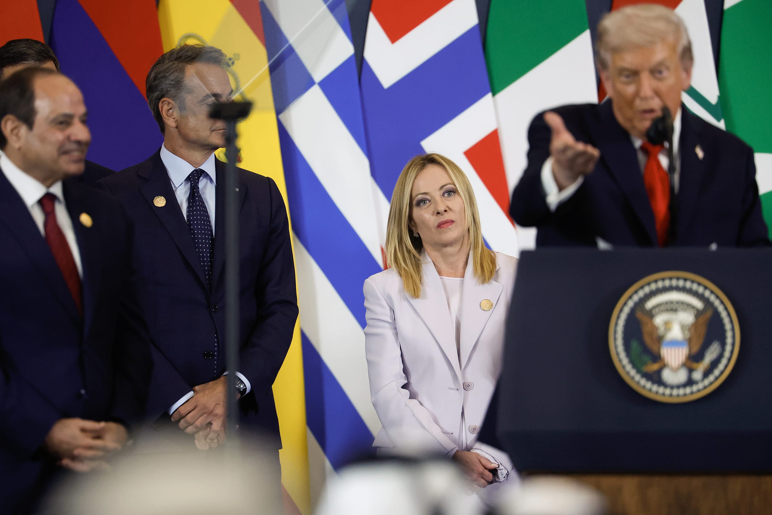 Donald Trump appears in soft focus at a podium in front of Giorgia Meloni.