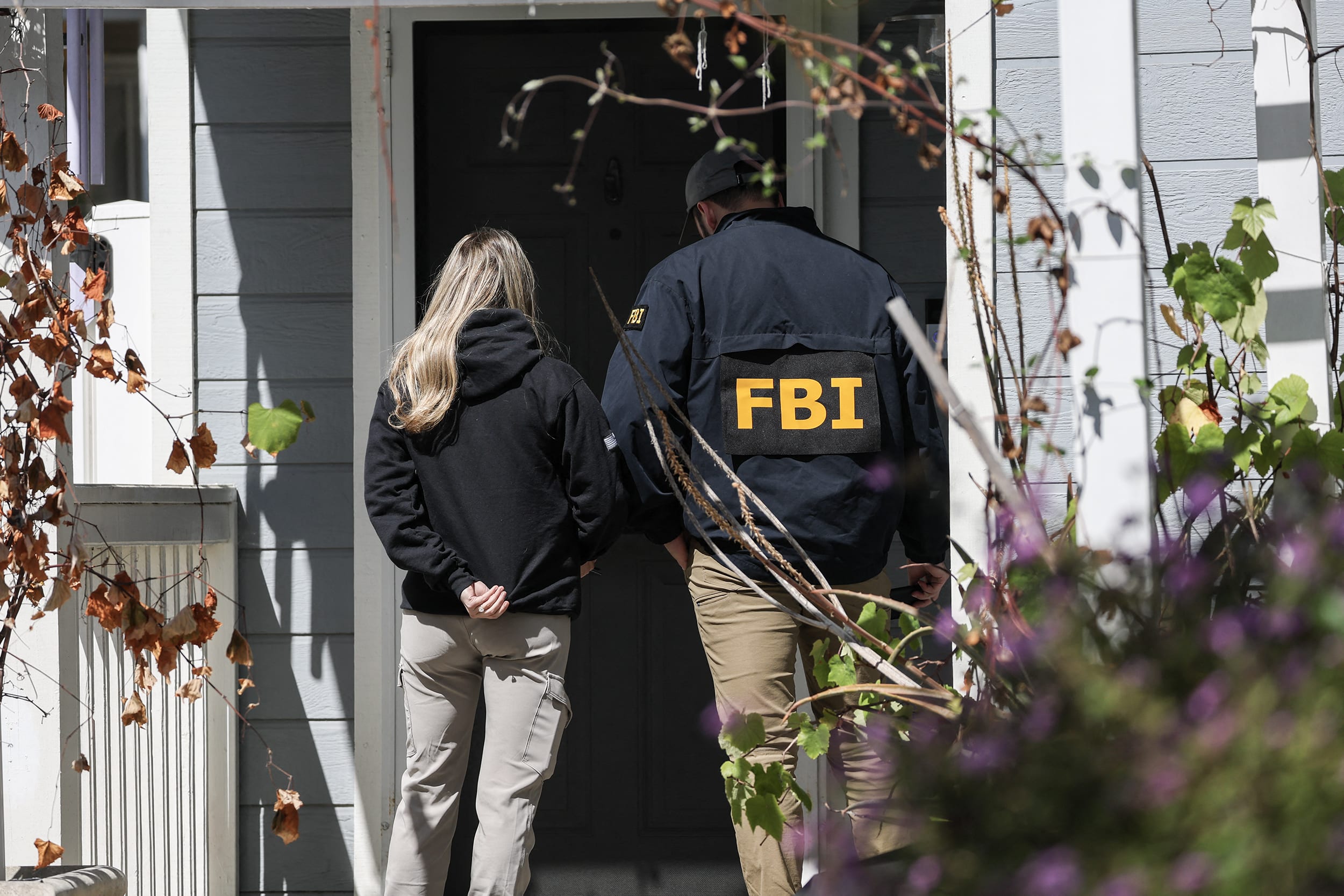 Two FBI field agents, one wearing a jacket with the agency's yellow lettering on its back, stand in front of a doorway to a residence.