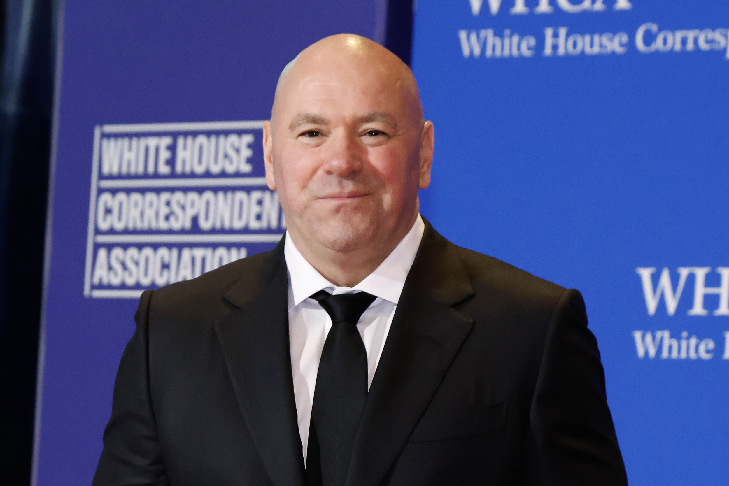 Dana White stands in front of a step-and-repeat background in a black suit.