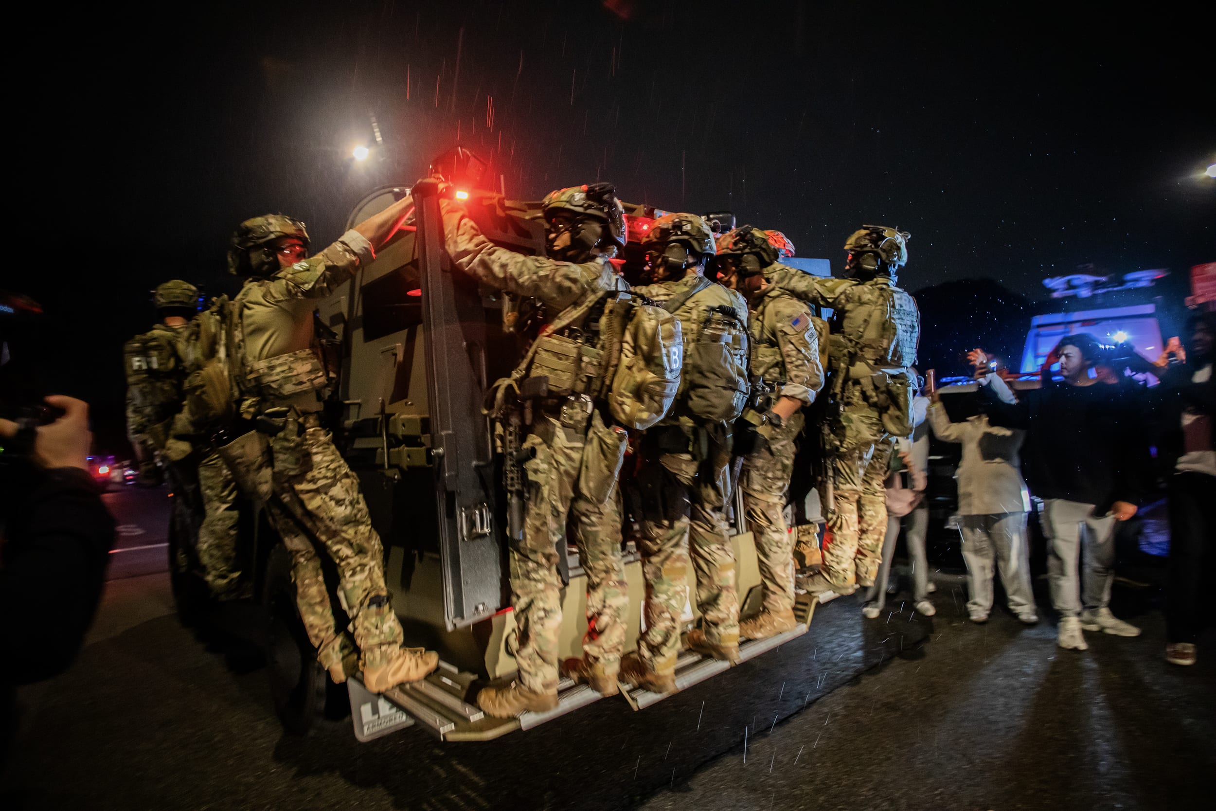 Numerous FBI agents wearing camouflage fatigues and helmets hold onto a SWAT vehicle as it moves down a city street in the rain. 