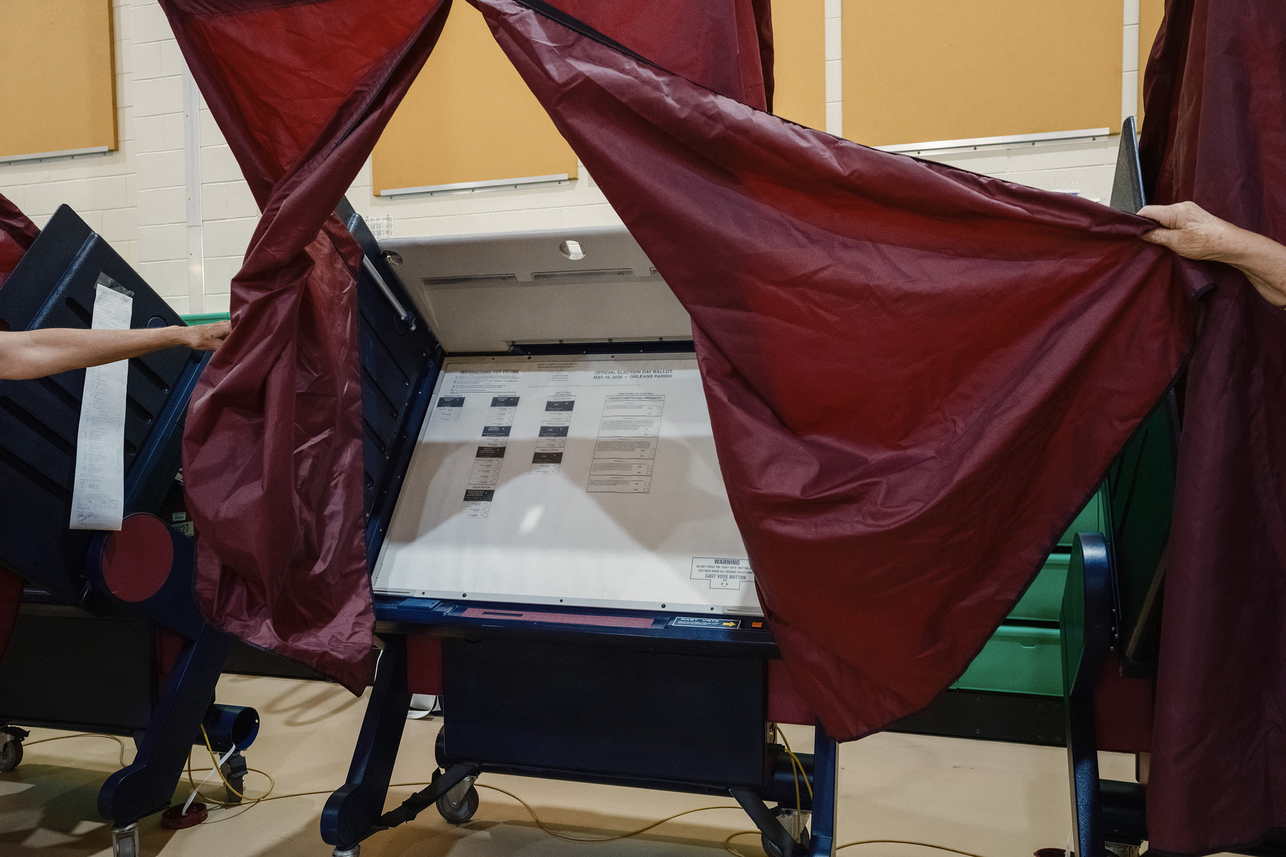 two outstretched arms pull back the red curtains to a ballot station inside a school gym.