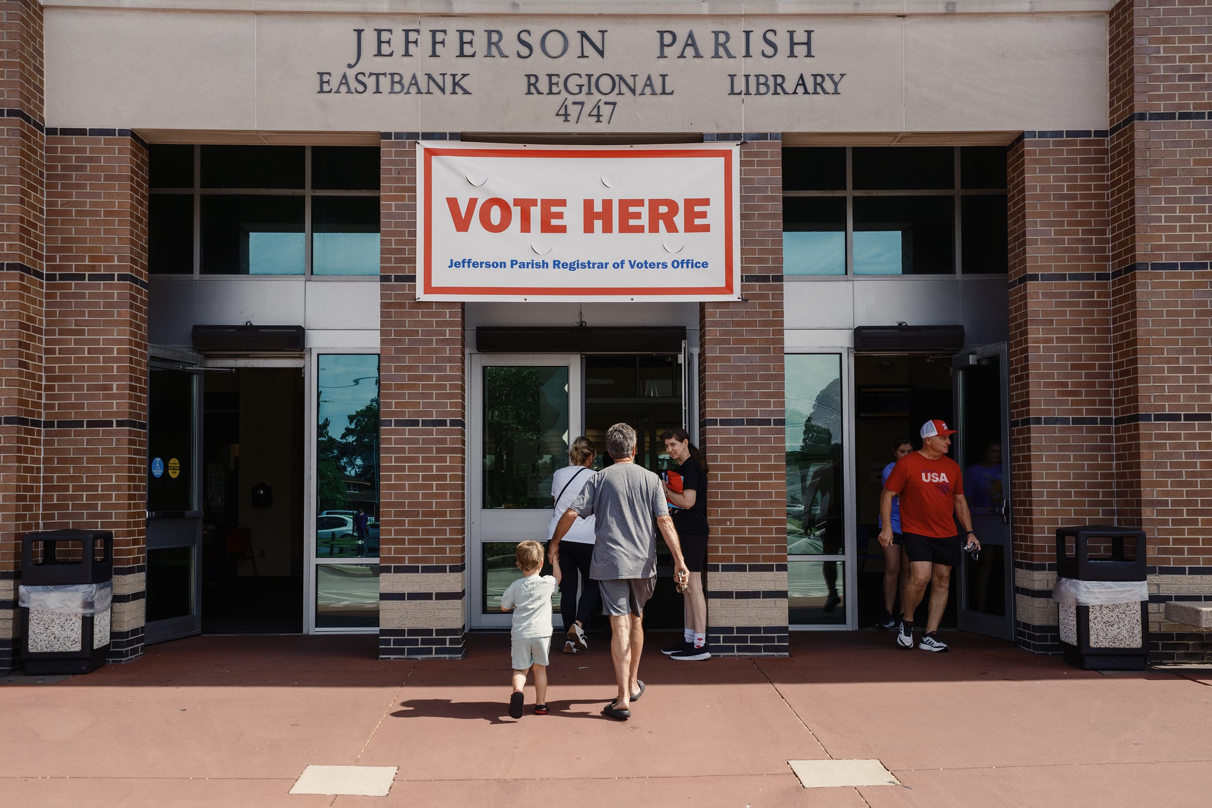 A VOTE HERE sign hung on the outside of a brick library building with people entering and exiting.