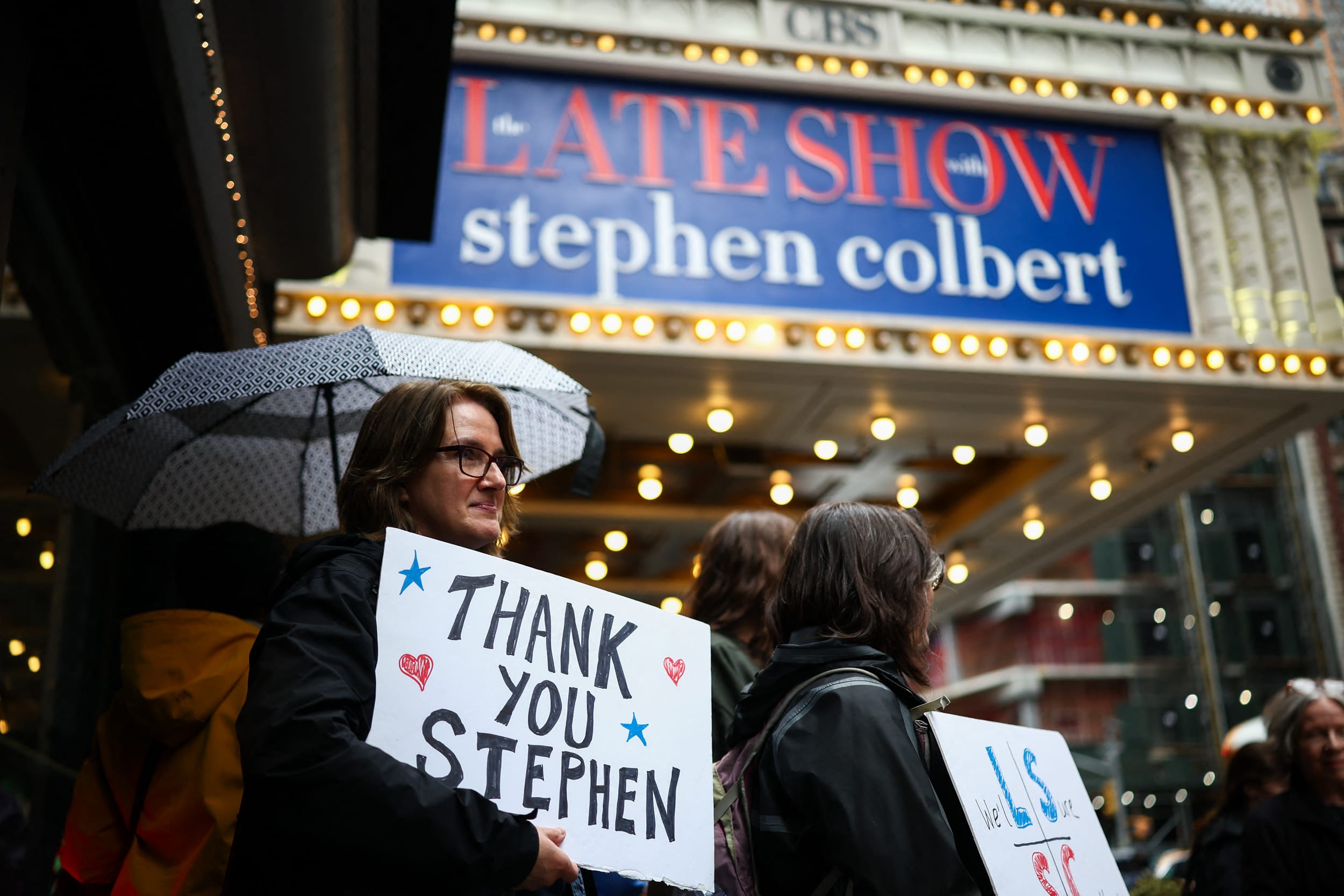 People hold signs supporting Stephen Colbert outside the Ed Sullivan Theater