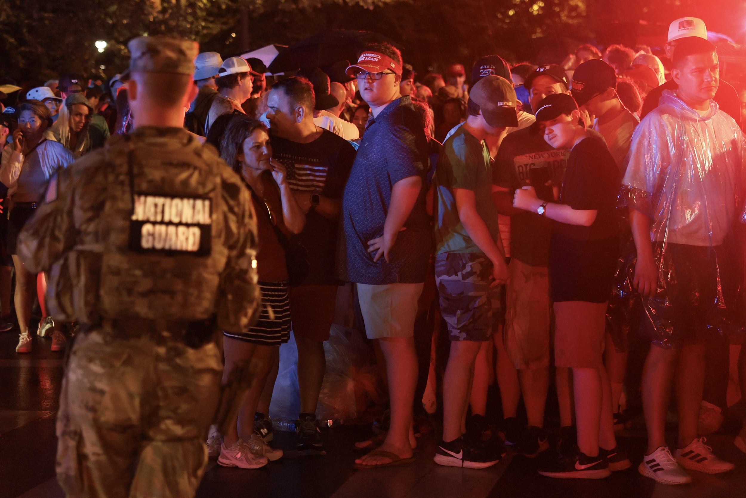 A thick crowd of people illuminated by red light stand behind a national guard soldier at night.