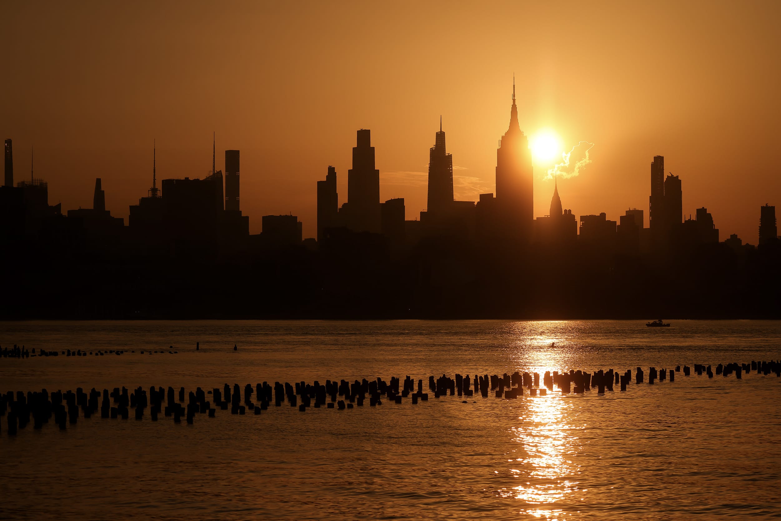 The sun rises over the Manhattan skyline and casts a long reflection along the water in front of it.