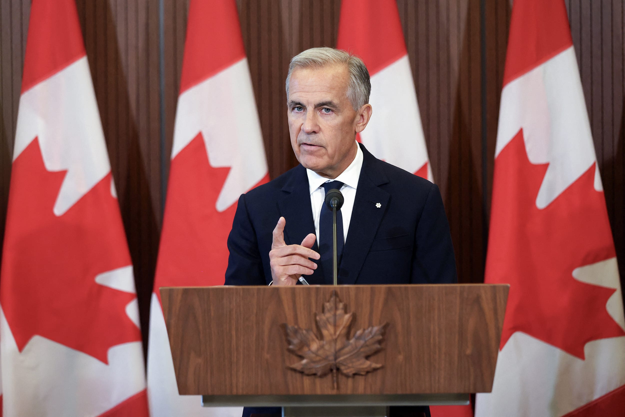 Mark Carney speaking at a wooden lectern in front of four Canadian flags.