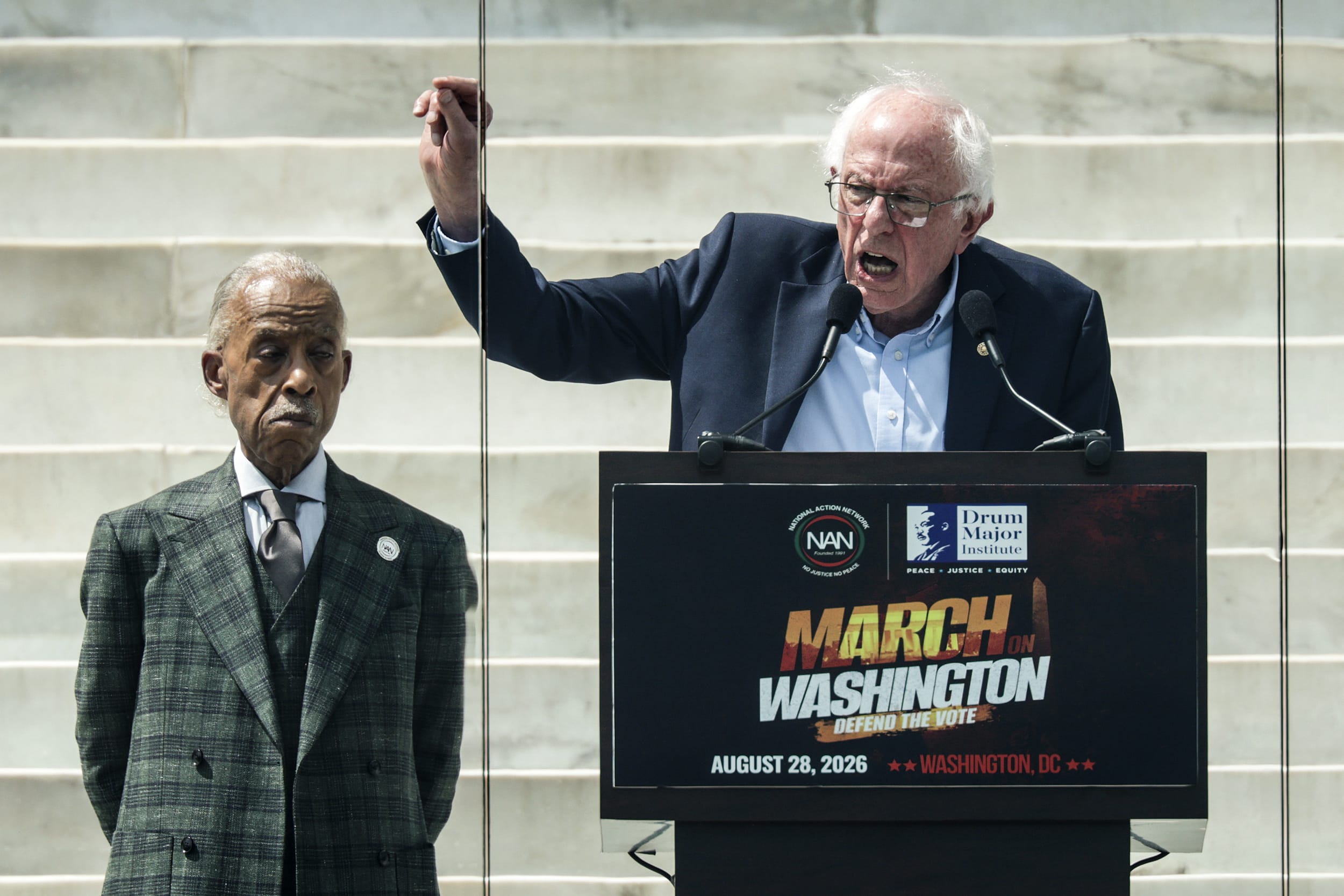 Bernie Sanders speaks at a lectern as Al Sharpton looks on behind him.
