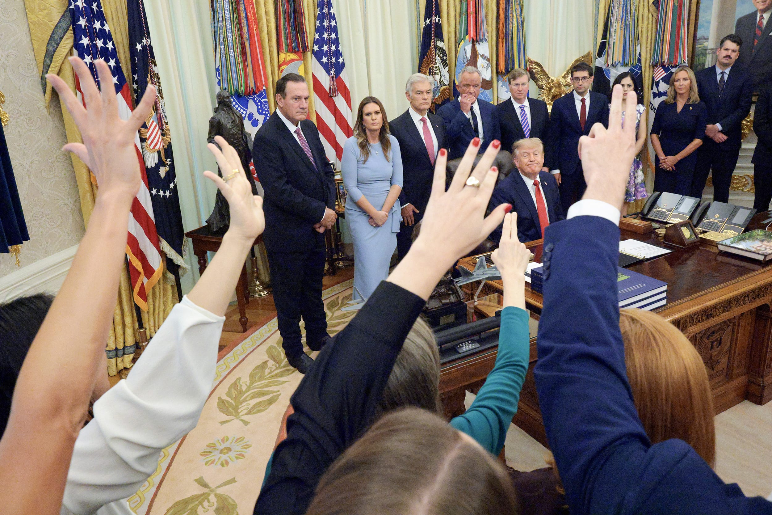 Reporters raise their hands in front of Donald Trump as he's seated at his desk in the Oval Office.