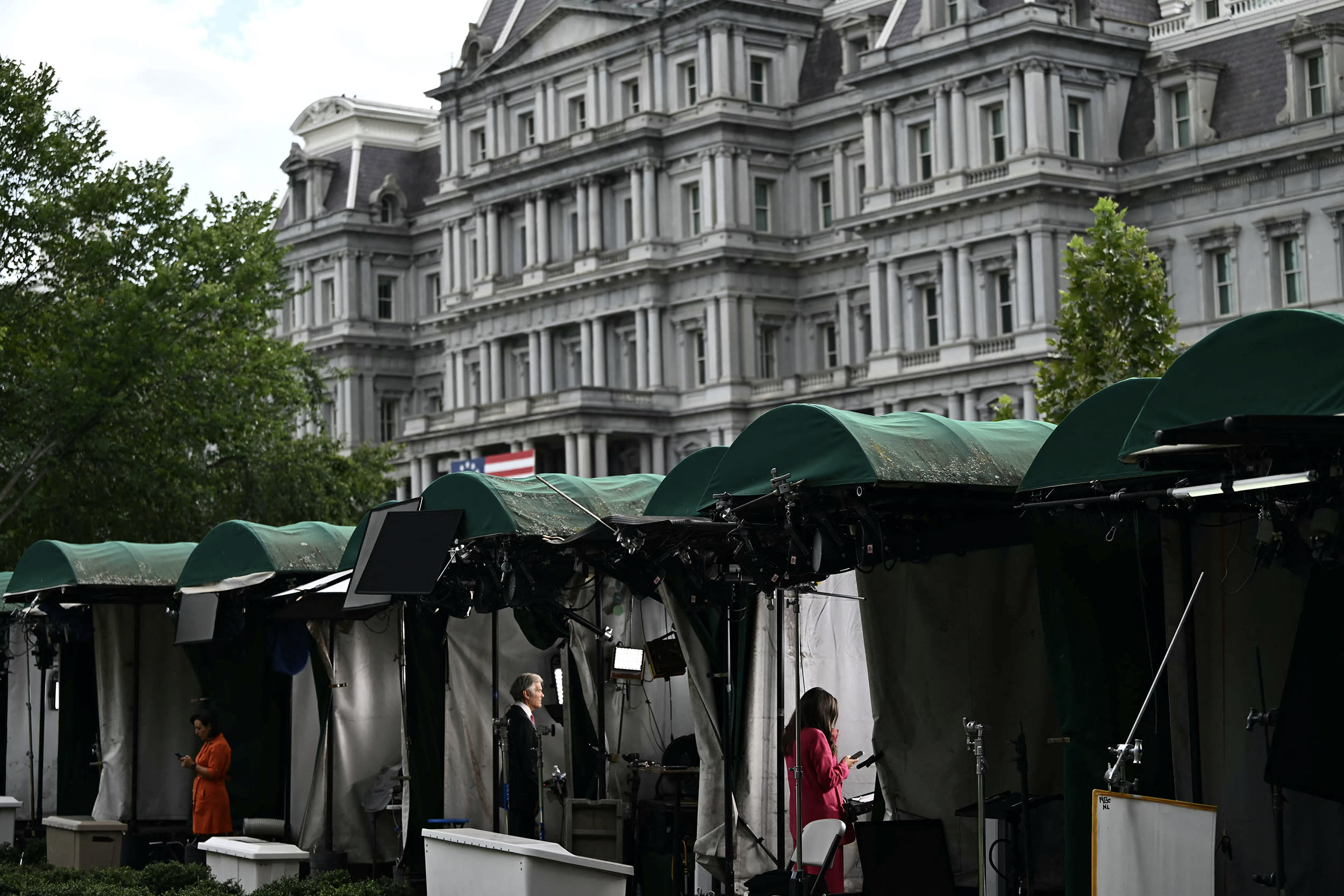 TV broadcast reporters stand inside green-roofed tents on the White House lawn.