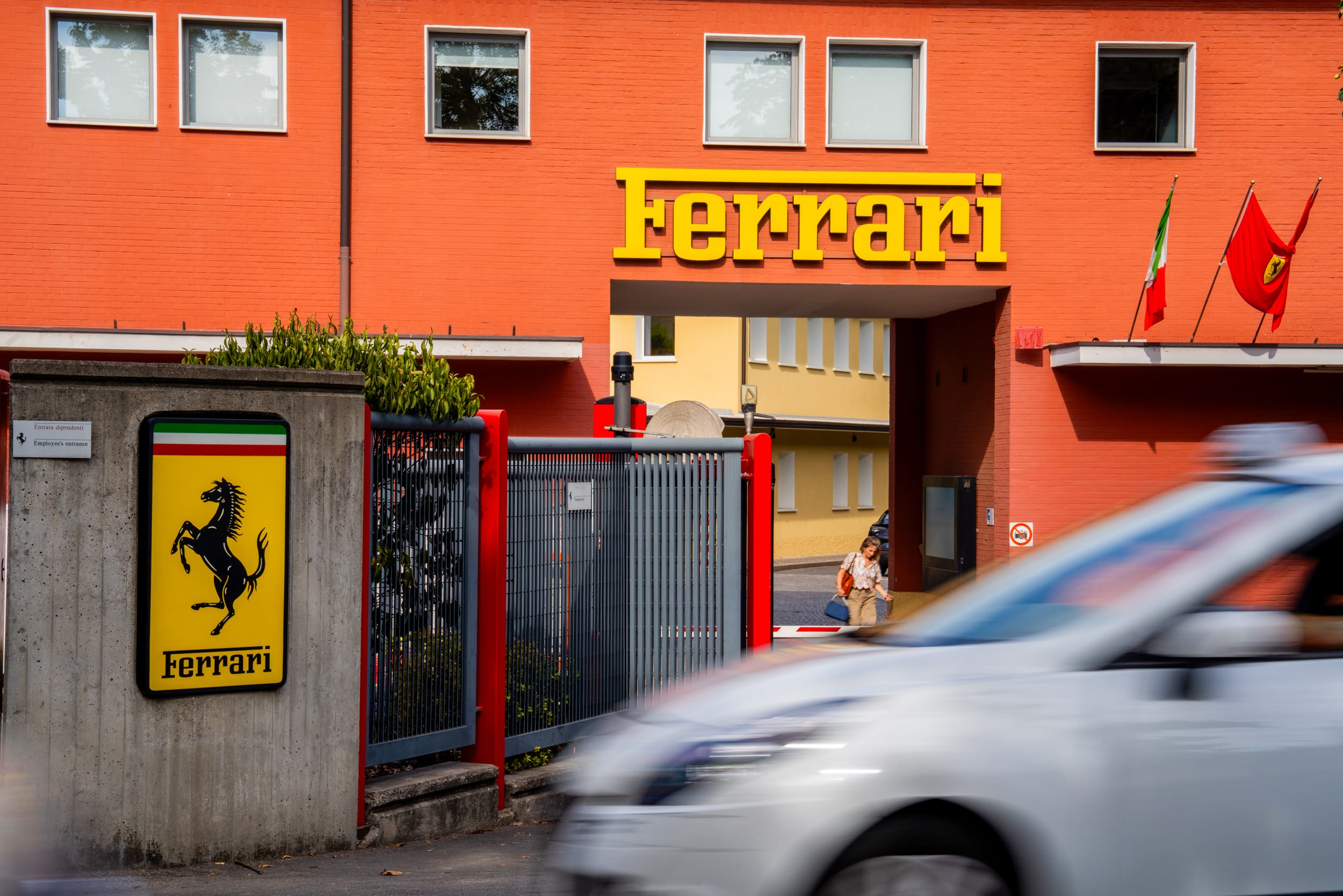 A detail view shows Ferrari factory signage, a yellow shield bearing the black prancing horse and Italian tricolor stripes, mounted on a concrete wall.