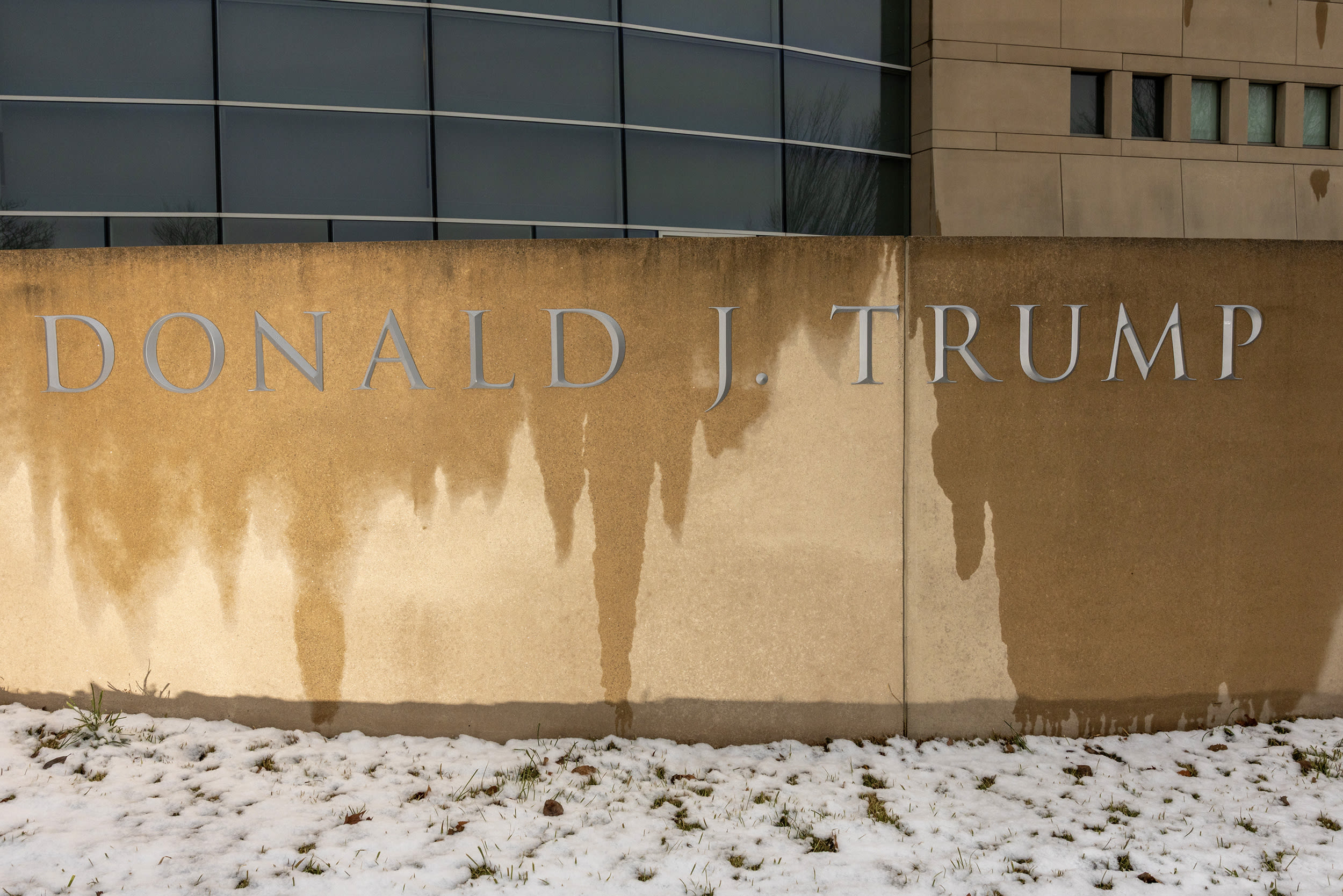 DONALD J. TRUMP etched into a wet stone facade, with snow covering the grass beneath it.