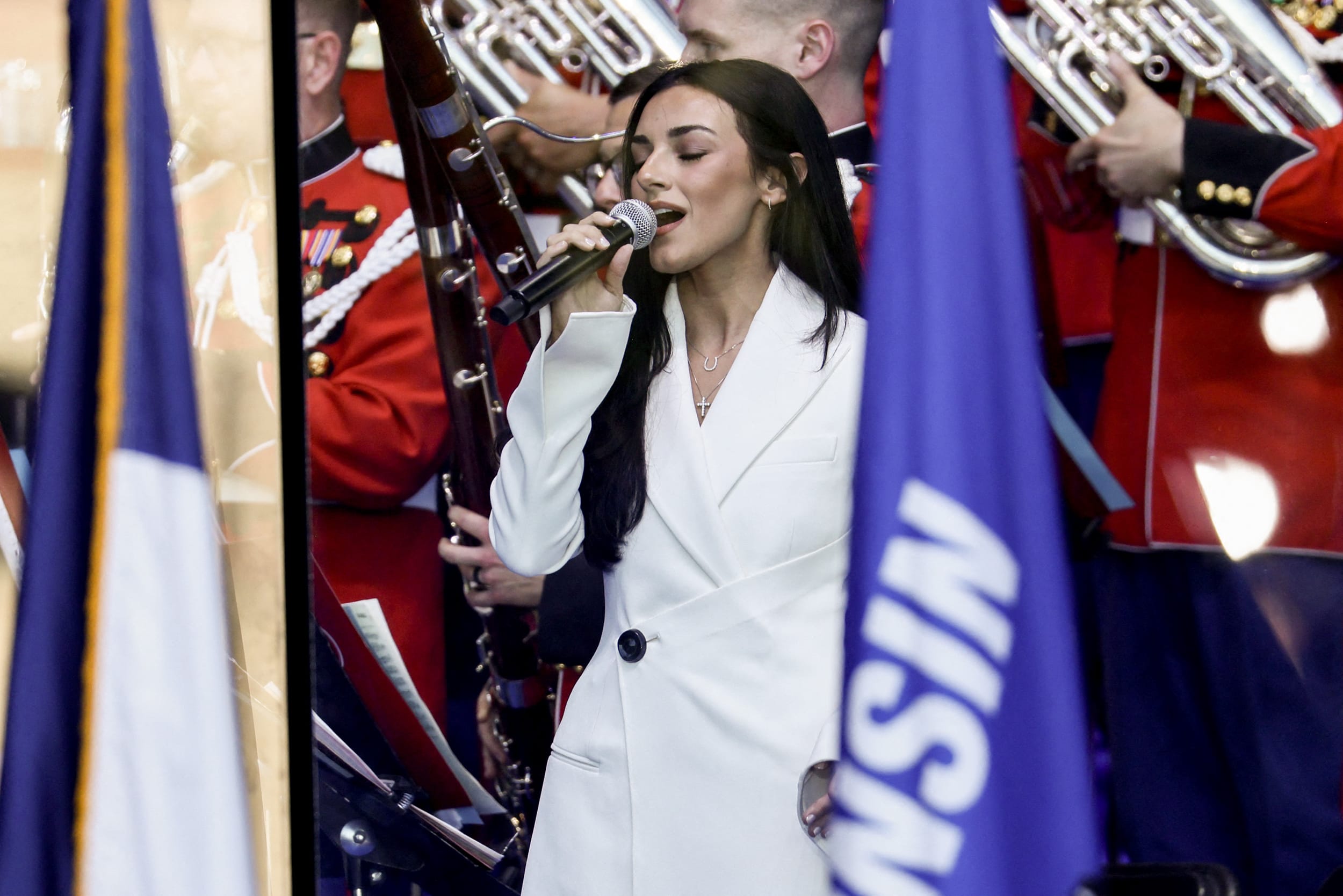 Alexis Wilkins singing into a wireless microphone in front of a marching band.