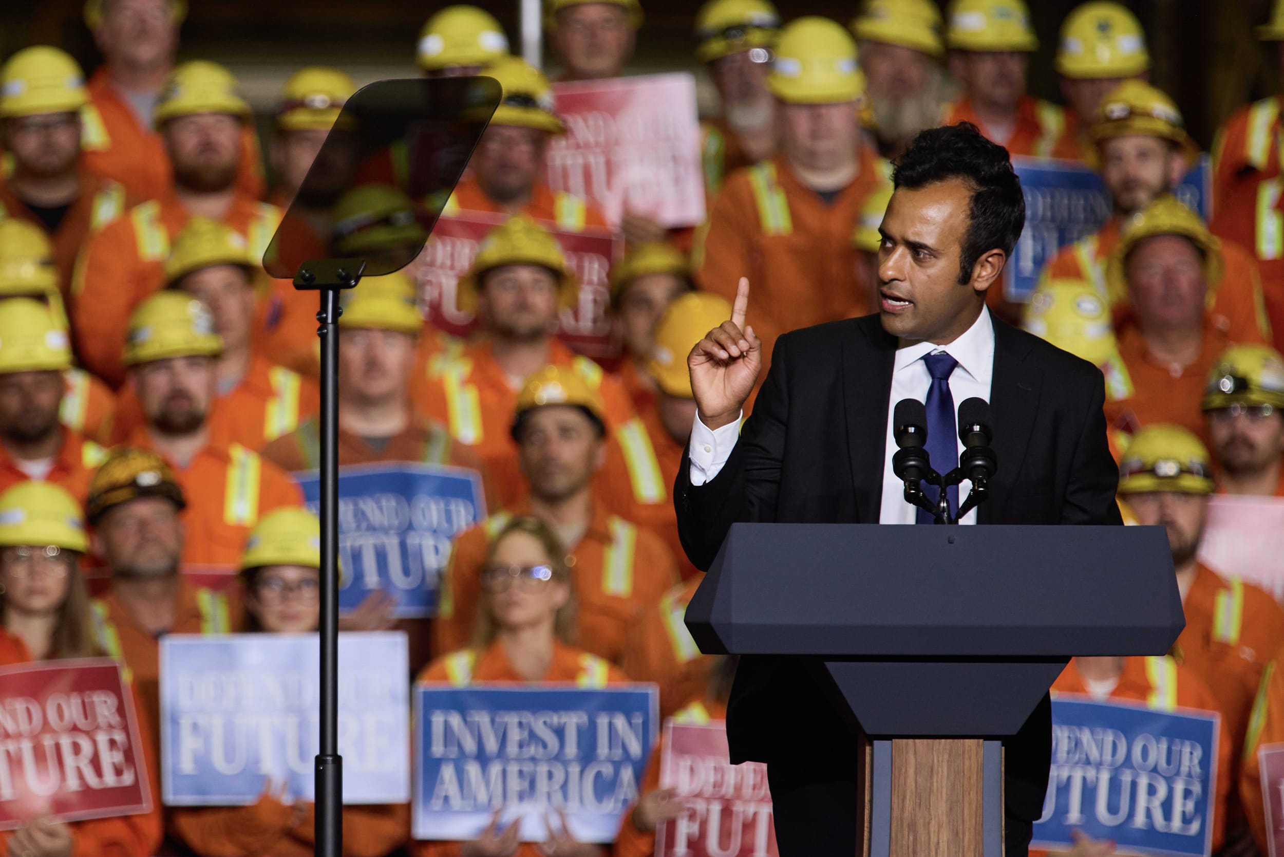 Vivek Ramaswamy speaking at a lectern in front of a riser full of people in orange vests and helmets.