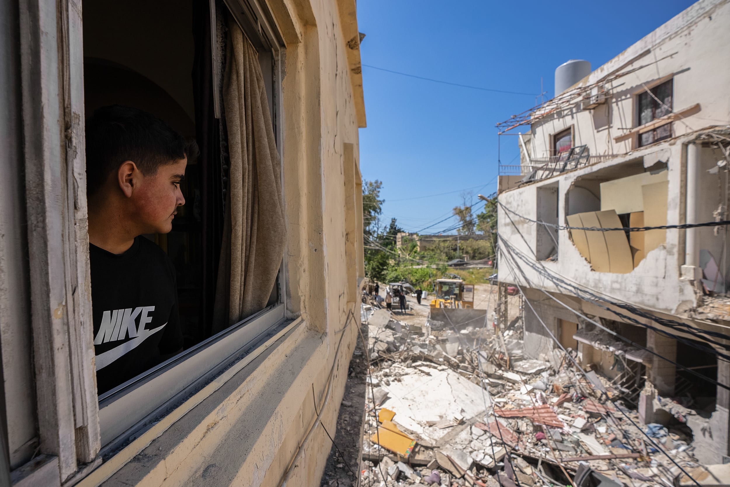 A young man wearing a black Nike t-shirt looks out a window toward rubble lying on the ground next to his building.