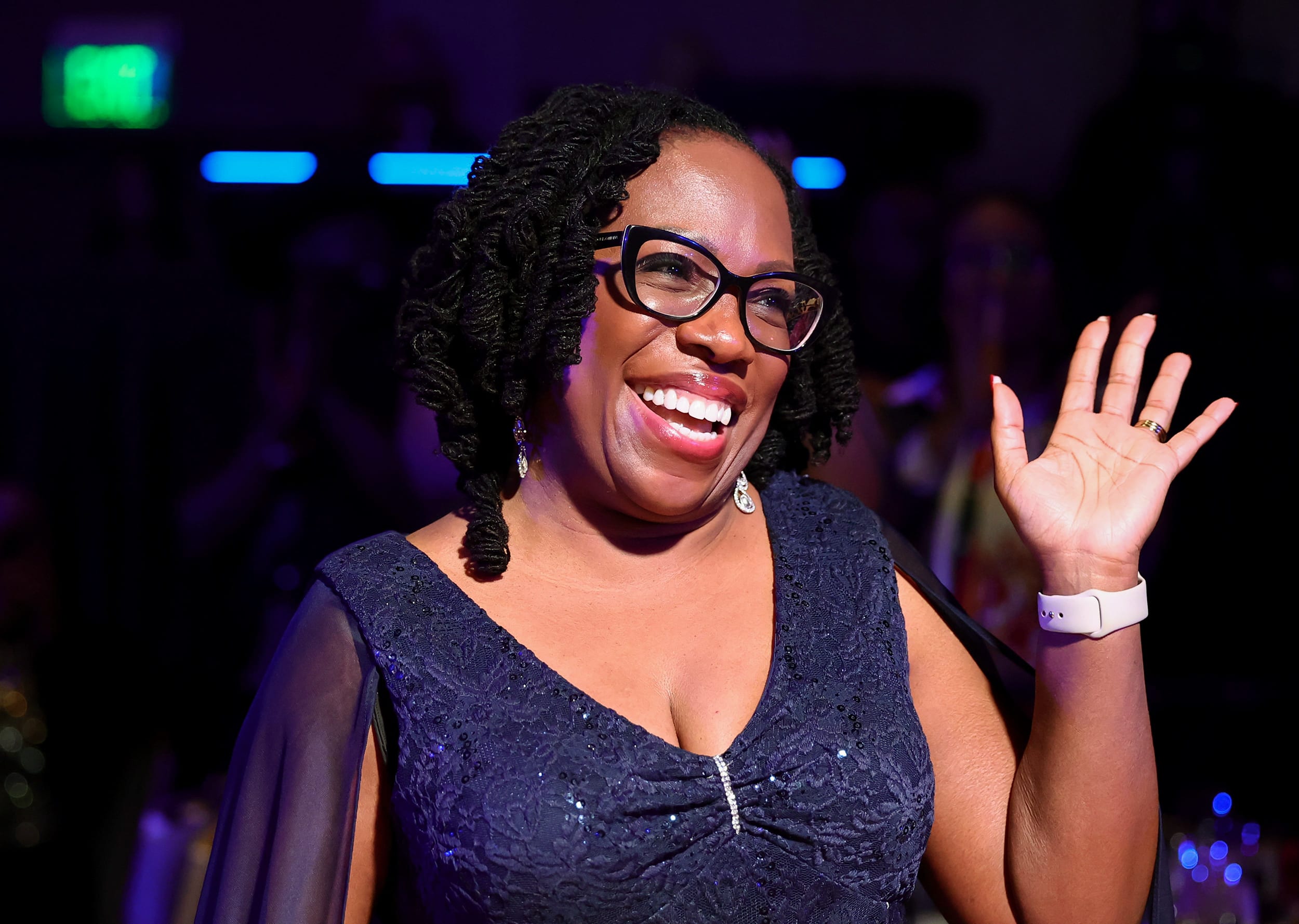 Ketanji Brown Jackson waves her left hand and smiles broadly inside a dimly-lit room with party lighting.