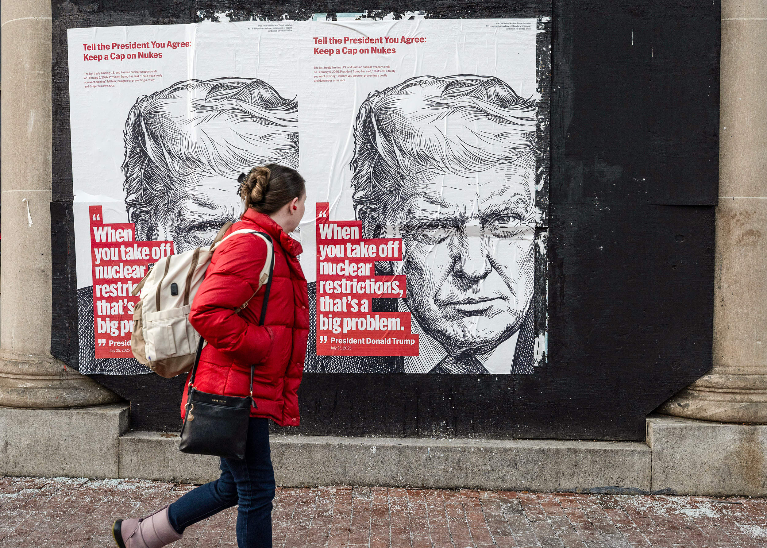 A woman in a red quilted jacket walks in front of two wheat-paste posters with an illustration of Donald Trump and a quote related to nuclear restrictions.