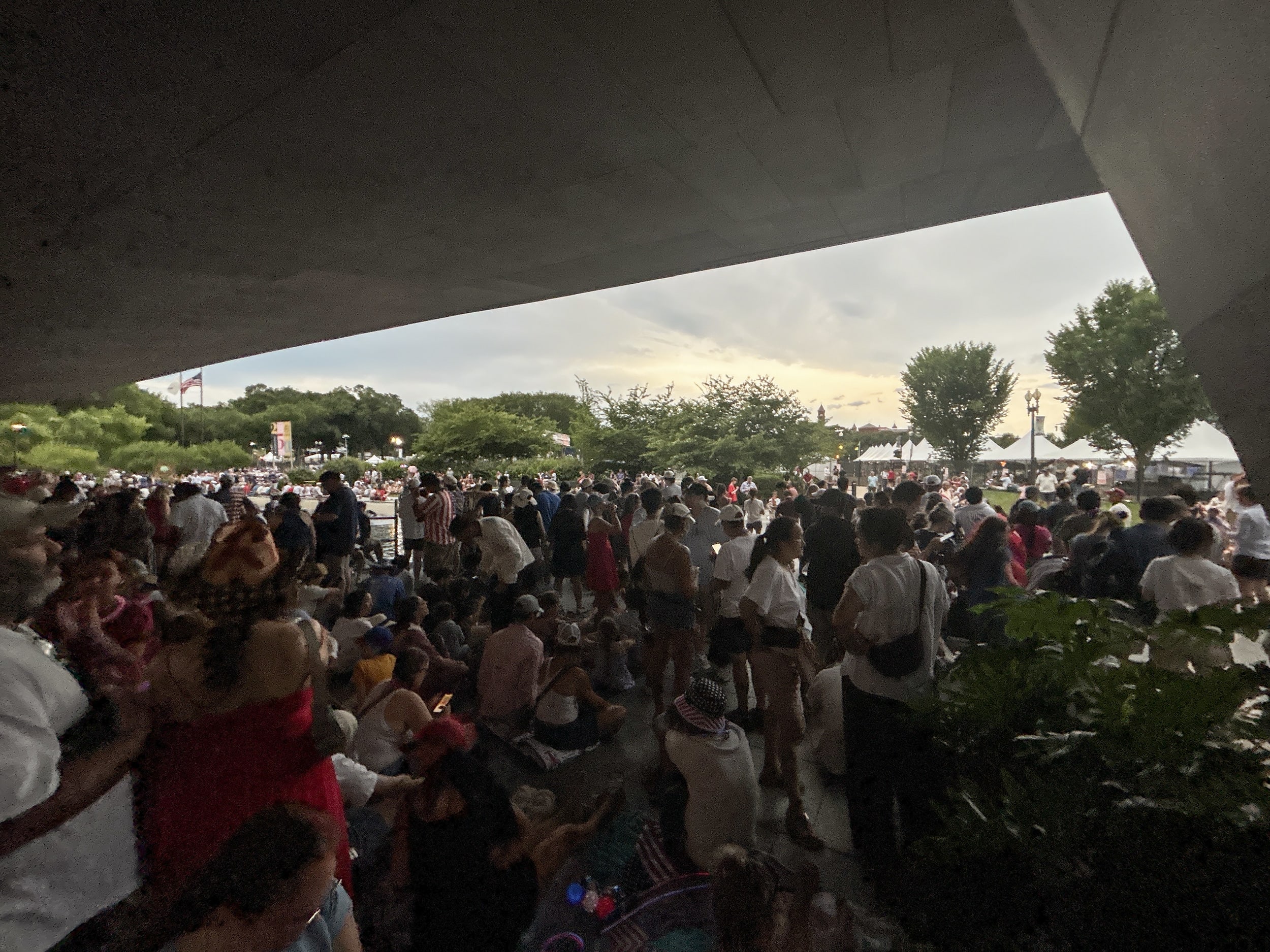 A large crowd of people sheltering from stormy skies under a concrete pavilion.