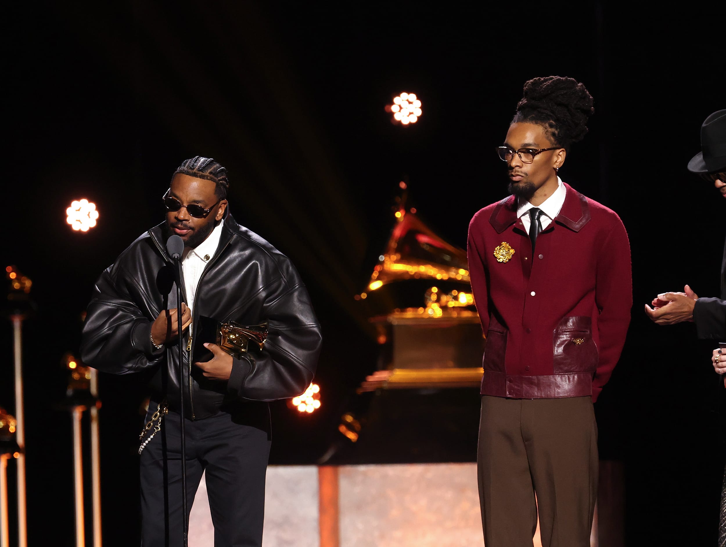 Sounwave holds a Grammy trophy while speaking into a microphone, wearing black sunglasses and a black leather jacket. Ruchaun Akers looks on from his left.
