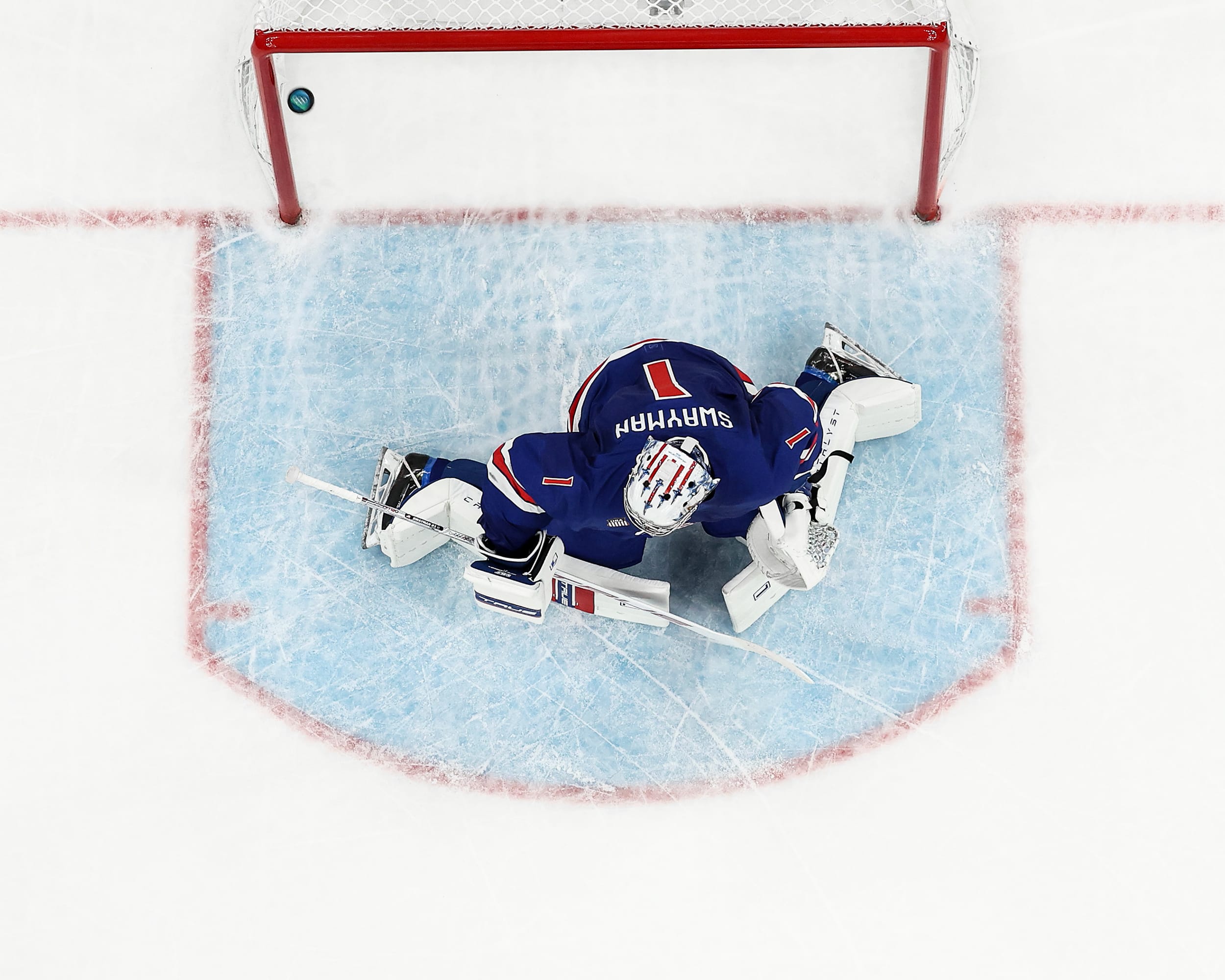 An overhead view of a goalie on the ice, in front of a puck that has drifted past the goal line and into the net.