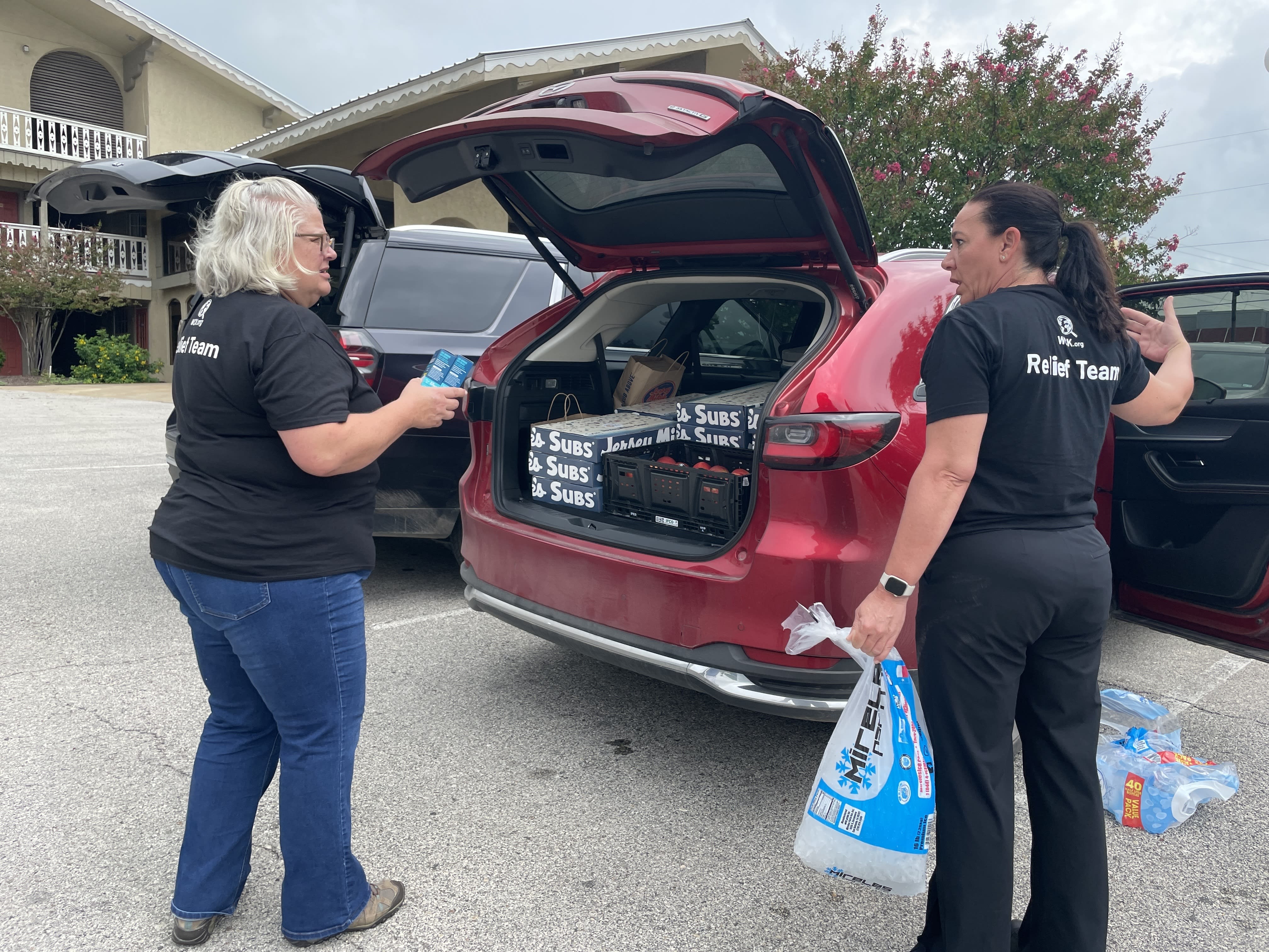 Revonda Kirby, left, and another person stand in front of the trunk of a car in a parking lot, holding bags of ice and other items