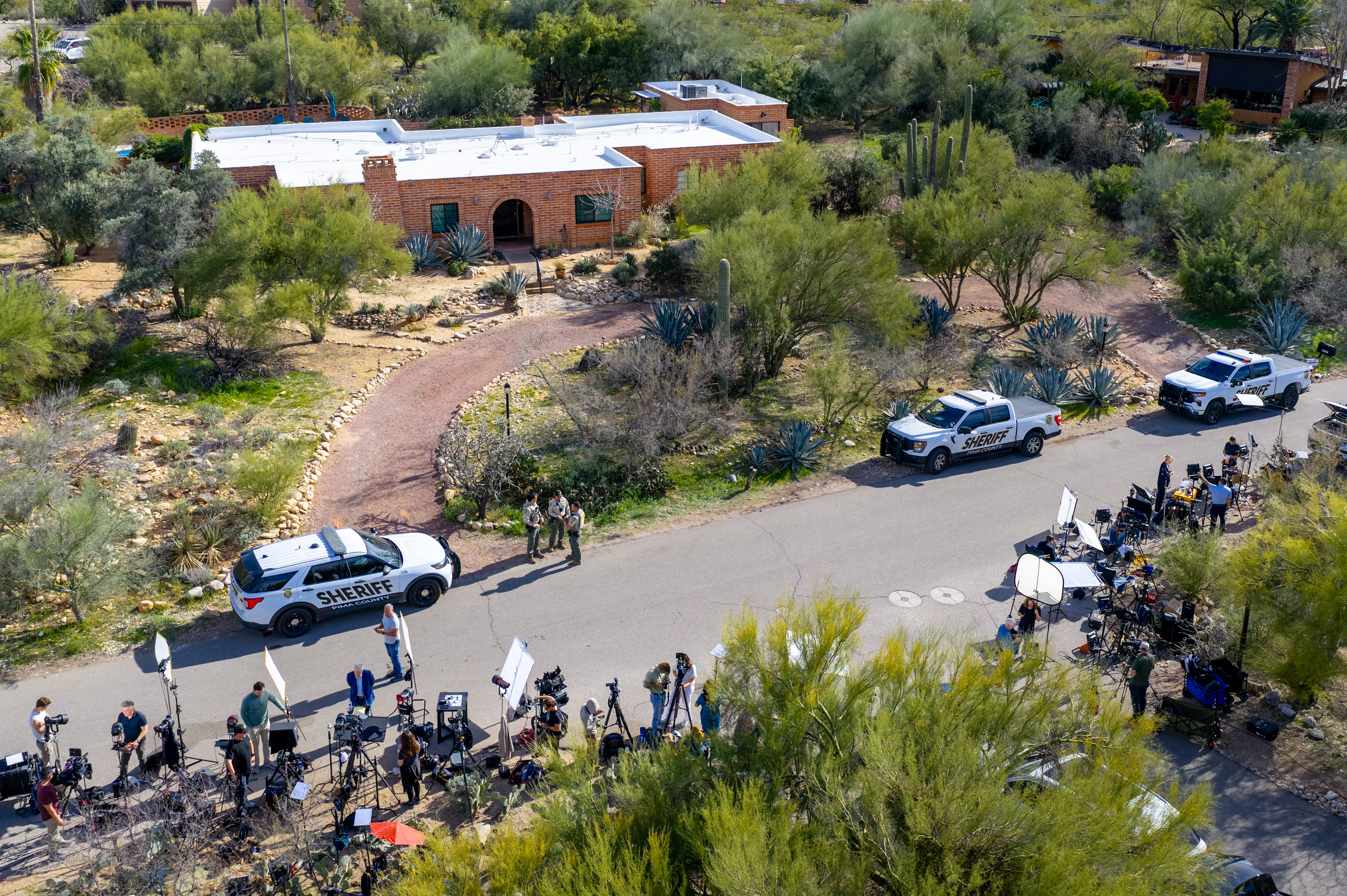 An aerial view of reporters and law enforcement officials gathered in the road in front of a single story brick home in a desert landscape.