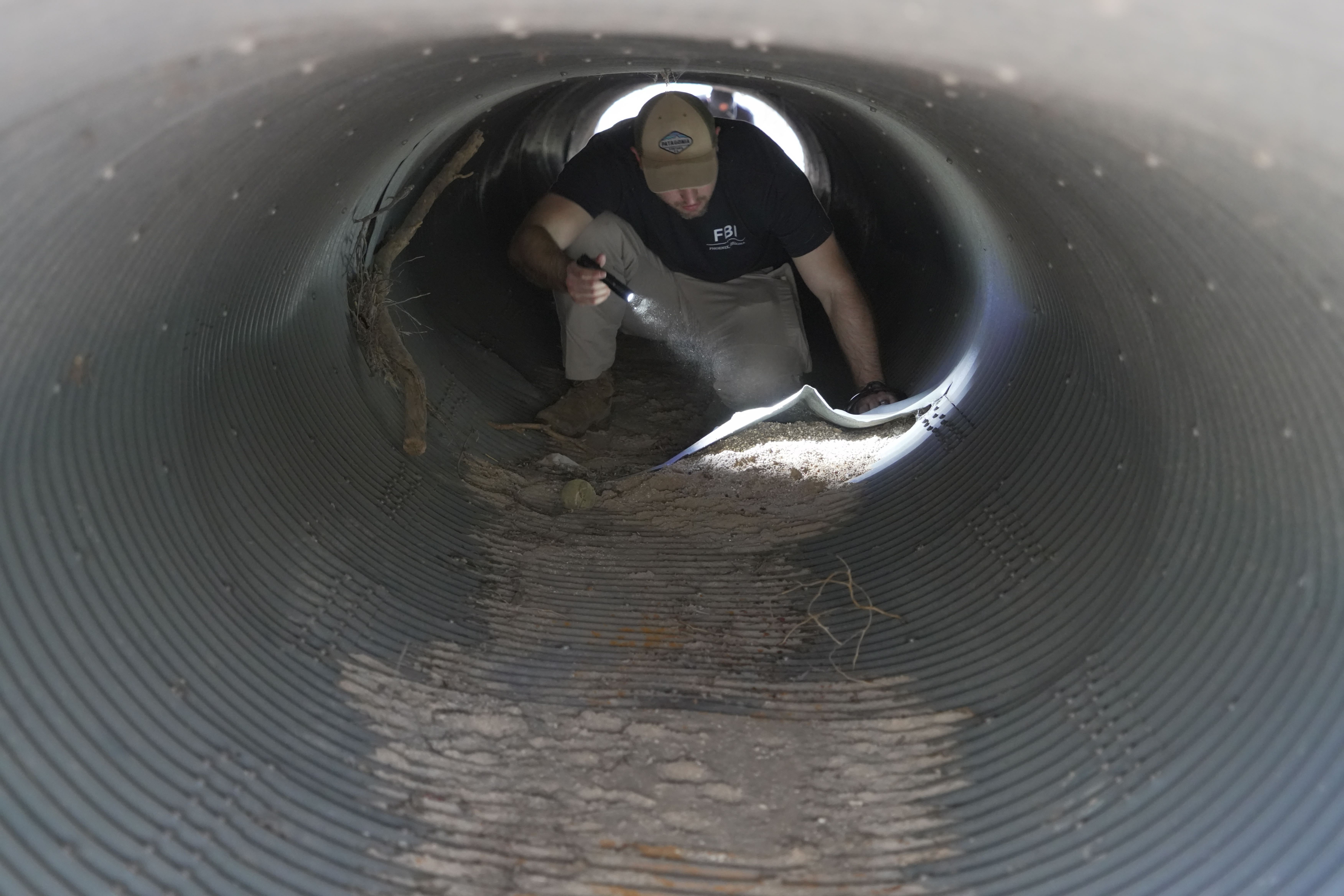 A man wearing an FBI t-shirt and holding a flashlight crawls on his hands and knees in a metal culvert.