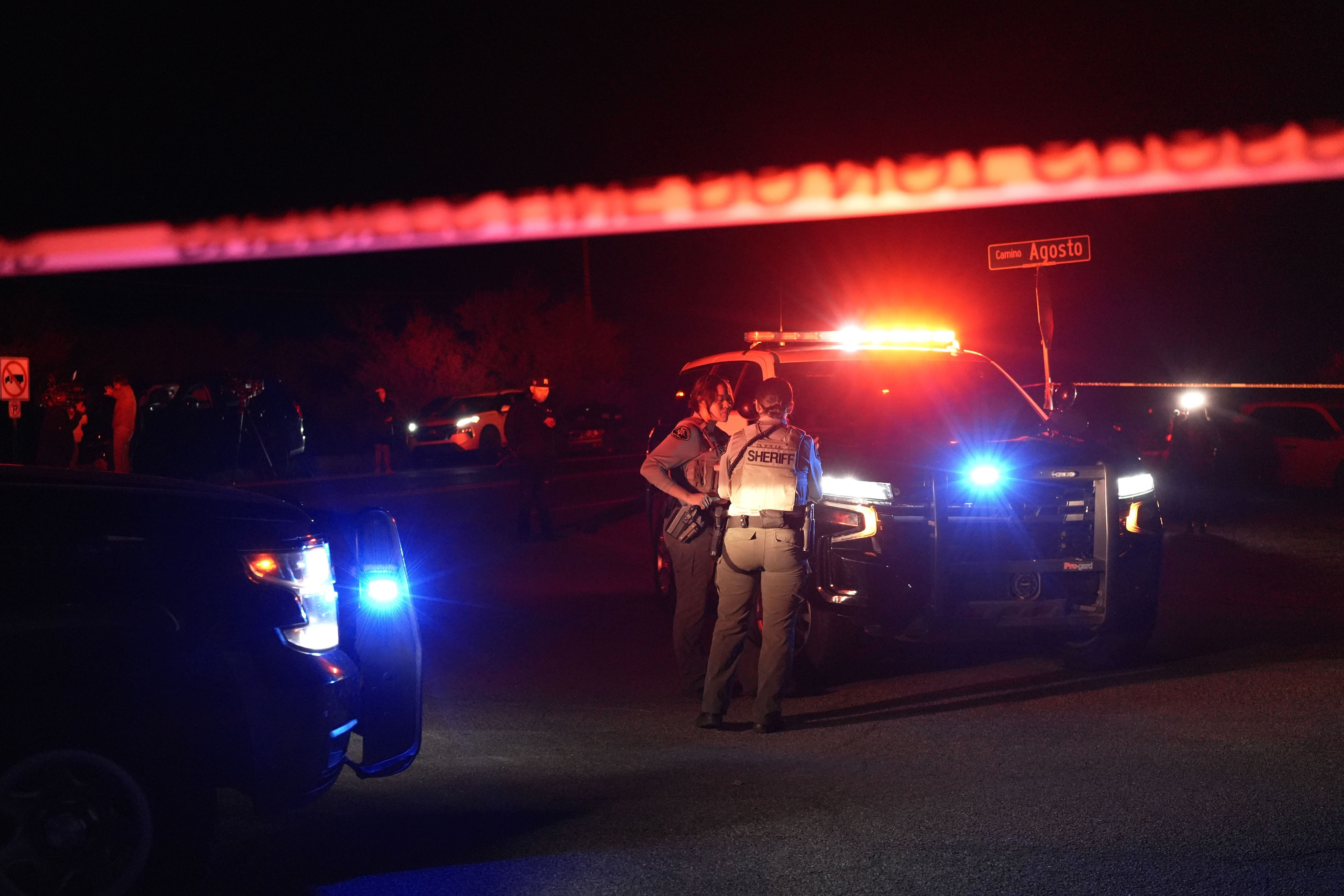 Two law enforcement officials stand in front of the flashing blue and red lights of a patrol vehicle, behind police tape that stretches across the frame.