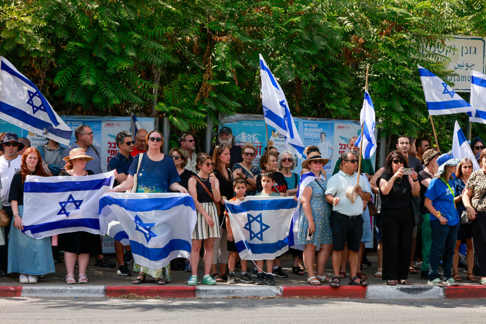 People line a sidwalk holding Israeli flags