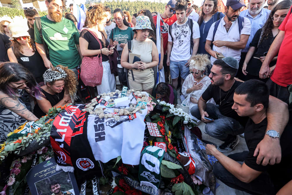 Mourners gather by a grave adorned with photos, rocks, and flowers