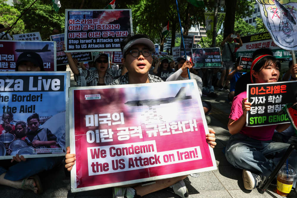 Protestors sit on the sidewalk outside and hold signs against U.S. and Israel attacks on Iran
