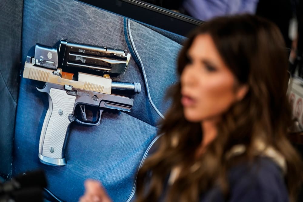 A photo of a gun is displayed on a screen next to a blurred Kristi Noem