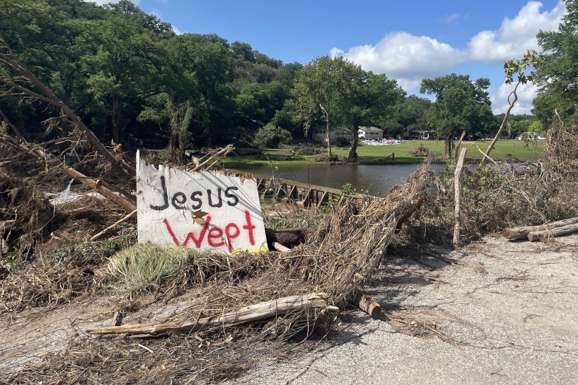 A sign that reads “Jesus Wept” sits propped up on debris across the river from Camp Mystic.