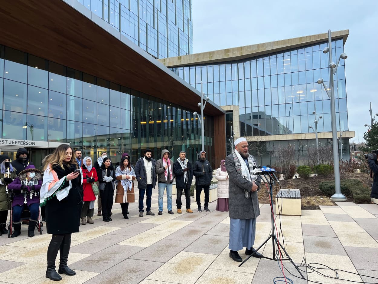 People gather for a vigil as a man prays at a set of microphones.