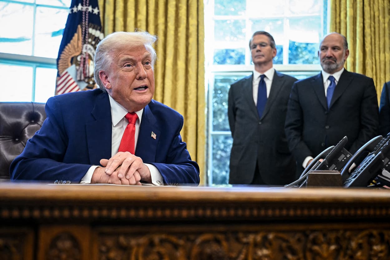 President Donald Trump, Treasury Secretary Scott Bessent, and Commerce Secretary Howard Lutnick inside of the Oval Office, Trump is seated at a desk, the other two are standing behind him