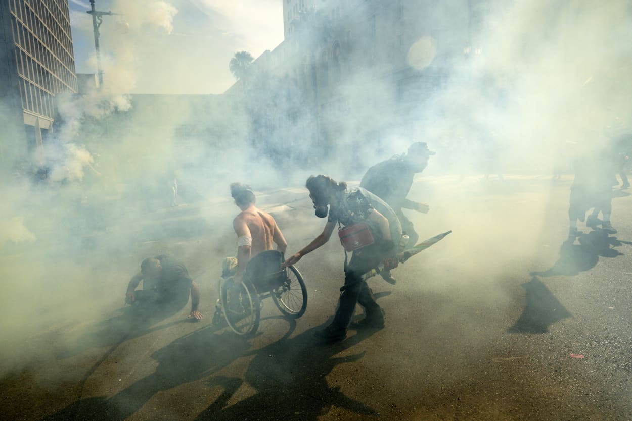 A man in a wheelchair and others move through tear gas clouds.