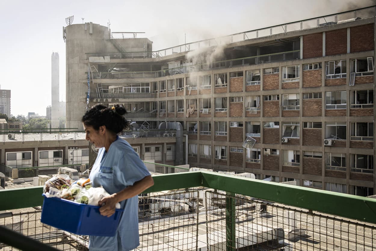 A nurse carries medical supplies past a building with smoke billowing out at Soroka Hospital
