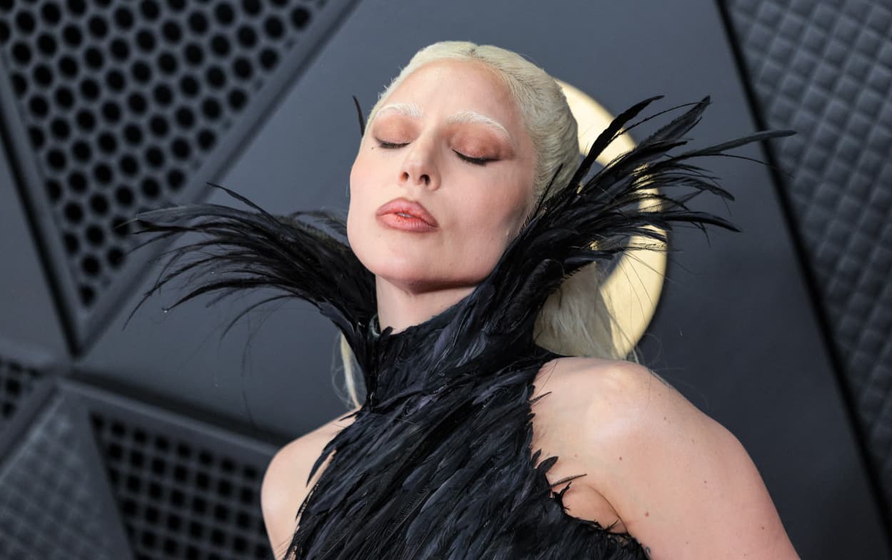 Lady Gaga, wearing a black feathered dress with a high collar, poses for a photo with her eyes closed. 