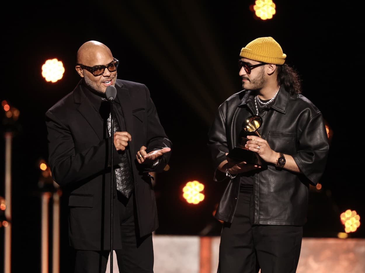 Israel Houghton speaks at a microphone while Aaron Moses stands next to him holding a Grammy award. 