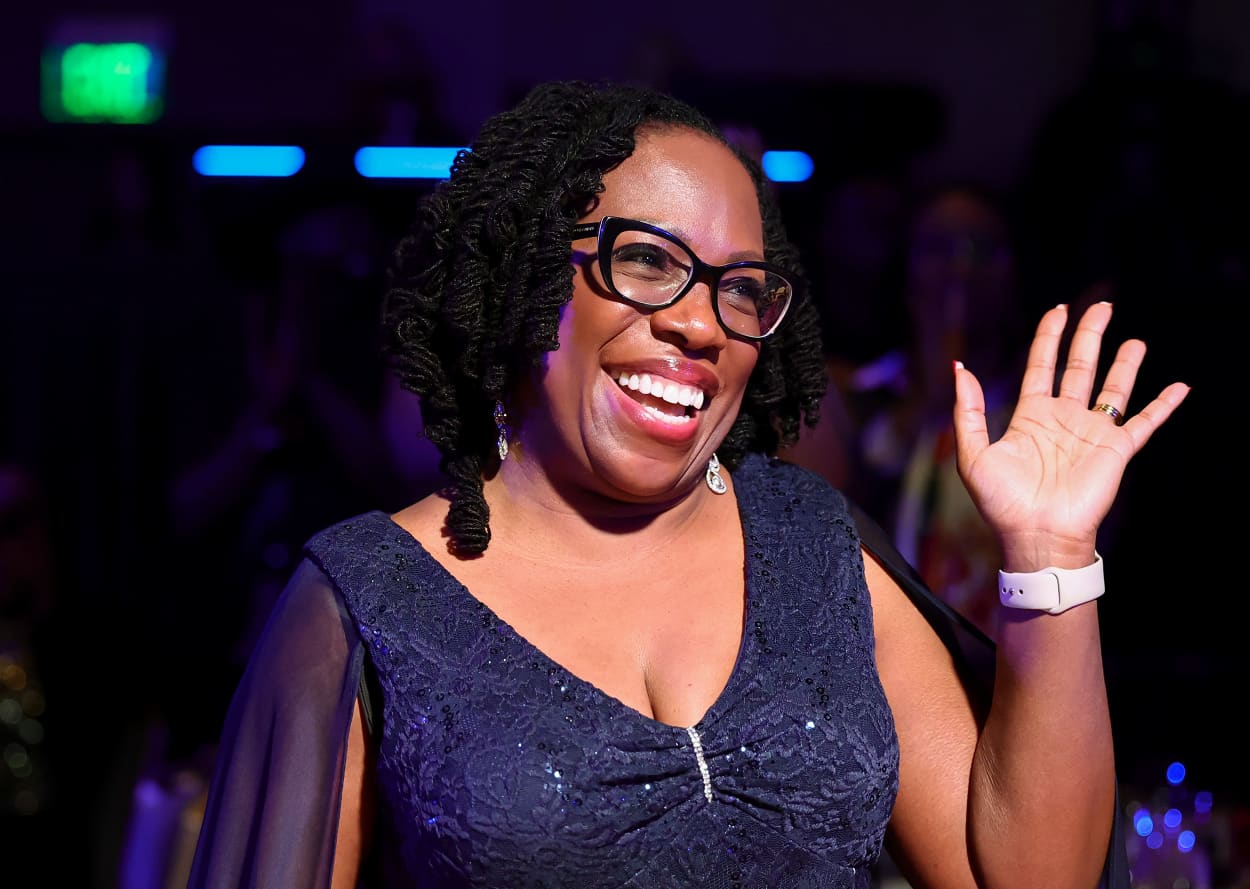Ketanji Brown Jackson waves her left hand and smiles broadly inside a dimly-lit room with party lighting.
