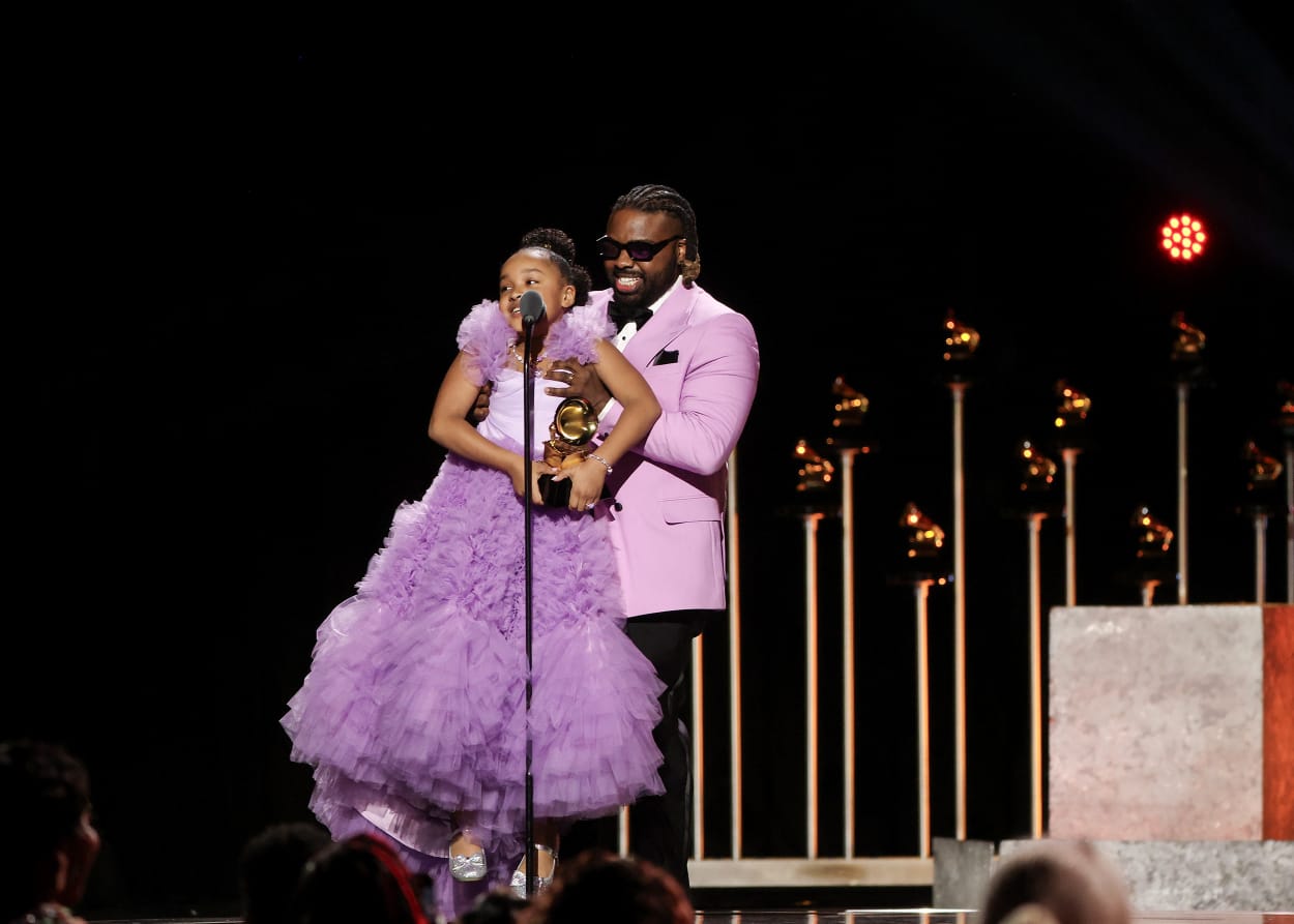 Fyutch holds Aura V up off the ground by her armpits so she can reach a microphone on stage at the Grammy awards. 